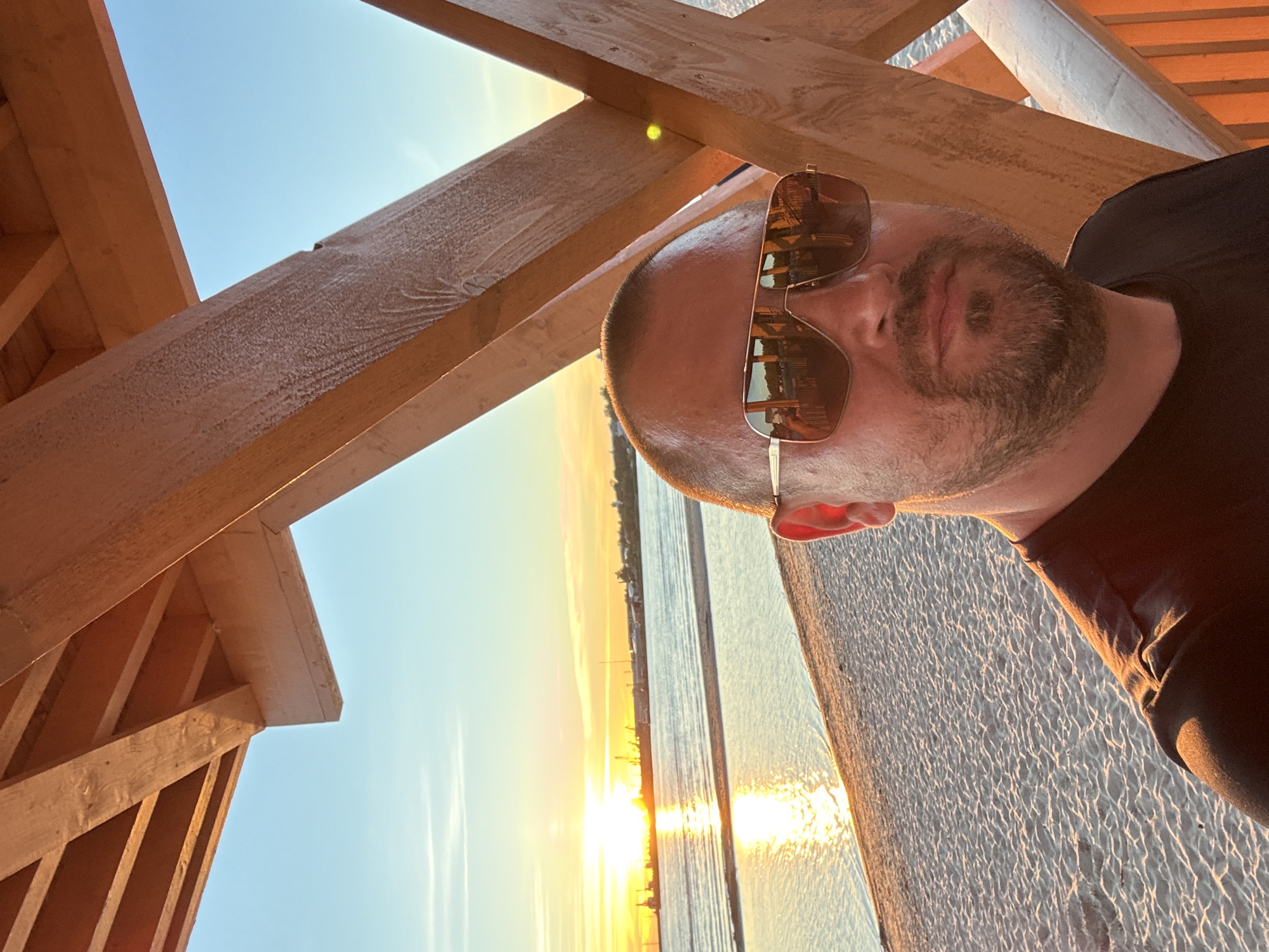 A man with sunglasses and a beard takes a selfie on a beach in Tallinn, Estonia. The sun is setting, casting a golden glow on the water and the sand. A wooden structure frames the scene, with its beams extending towards the sky. The man is wearing a black t-shirt. The beach is mostly empty, with a few boats visible in the distance on the water. The sky is a clear blue, with a few wispy clouds.