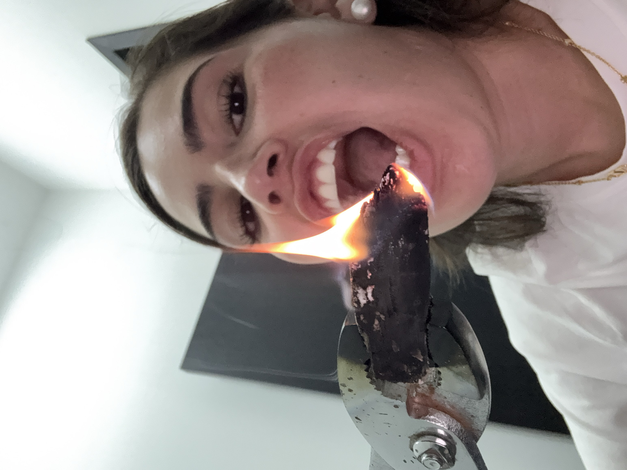 A low-angle shot captures a woman with dark hair, pearl earrings, and a gold necklace, tilting her head back with her mouth open. In the foreground, metal tongs hold a burning, dark-colored object, with flames reaching toward her open mouth. The background includes a glimpse of a dark square shape against a white wall. The location is not discernible from the provided image. No actions or interactions are taking place other than the positioning of the items in relation to each other.
