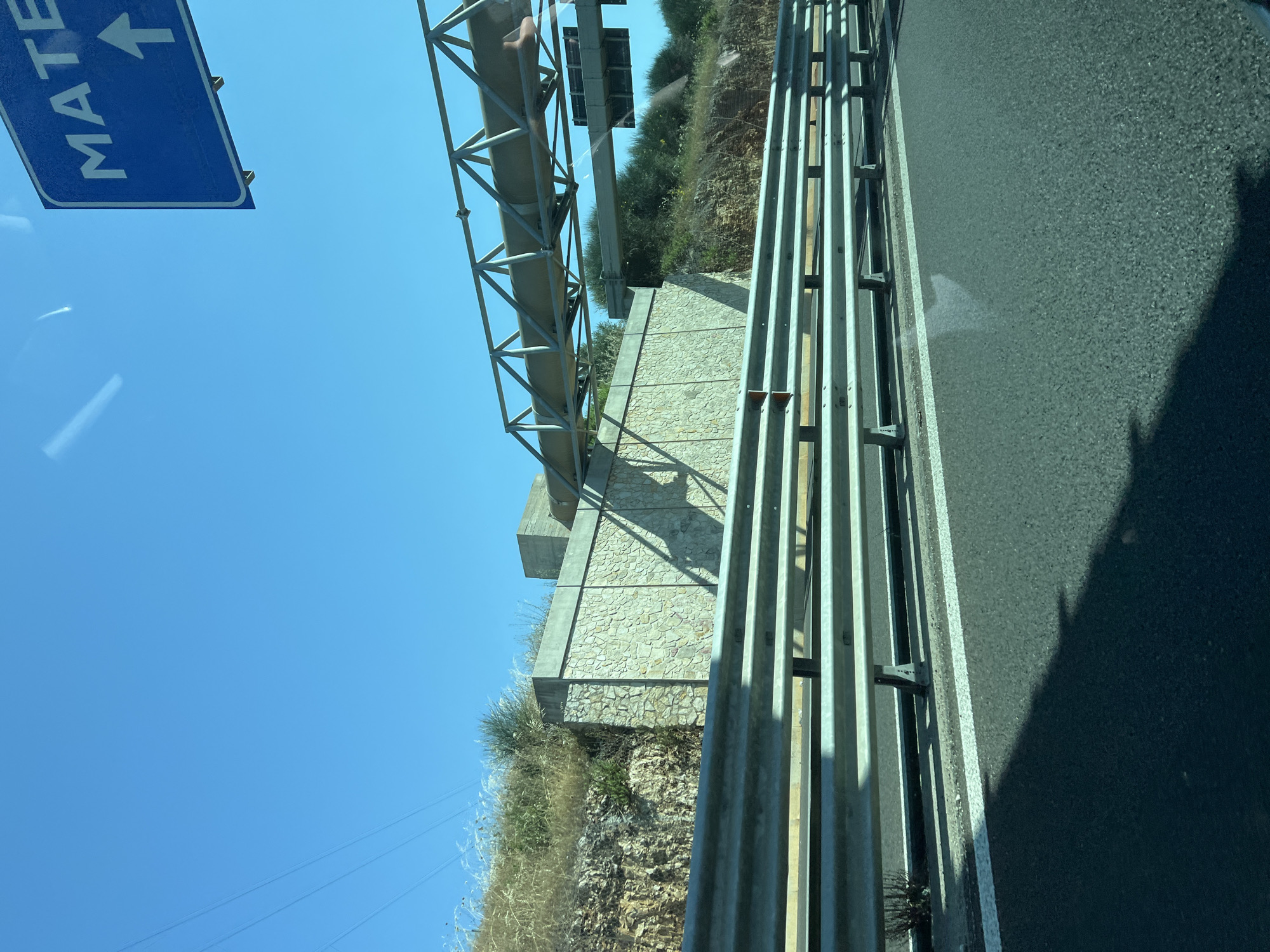 This image captures a bright, sunny daytime scene of a highway, assumed to be near Palo del Colle, Italy. In the immediate foreground, a multi-layered metal guardrail runs horizontally across the frame, separating the dark asphalt road from the adjacent terrain. The asphalt surface exhibits white lane markings on the right side, and distinct shadows are cast onto the road from the guardrail and the unseen vehicle from which the photograph was taken.
In the midground, beyond the guardrail, a rugged, dry grassy and scrub-covered hillside with exposed rocky sections rises on the left. A light-colored stone retaining wall, constructed from irregularly shaped blocks, supports a portion of this hillside. Above the retaining wall, another, smaller concrete structure is visible. Extending across the highway in the upper midground is a large metal truss structure, appearing to be an overhead gantry or bridge, supporting a large, fabric-wrapped pipeline or conduit. Further down the road, another, simpler overhead gantry supports dark, rectangular objects, likely road signs or electronic equipment.
The background is dominated by a clear, bright blue sky. In the upper left, power lines are faintly visible against the sky. On the upper right, a portion of a blue road sign is visible, displaying "MATE" (likely indicating Matera) and an upward-pointing white arrow, suggesting a straight-ahead direction for the destination. No people are visible in the scene. The intense sunlight creates stark shadows, indicating a high sun position.