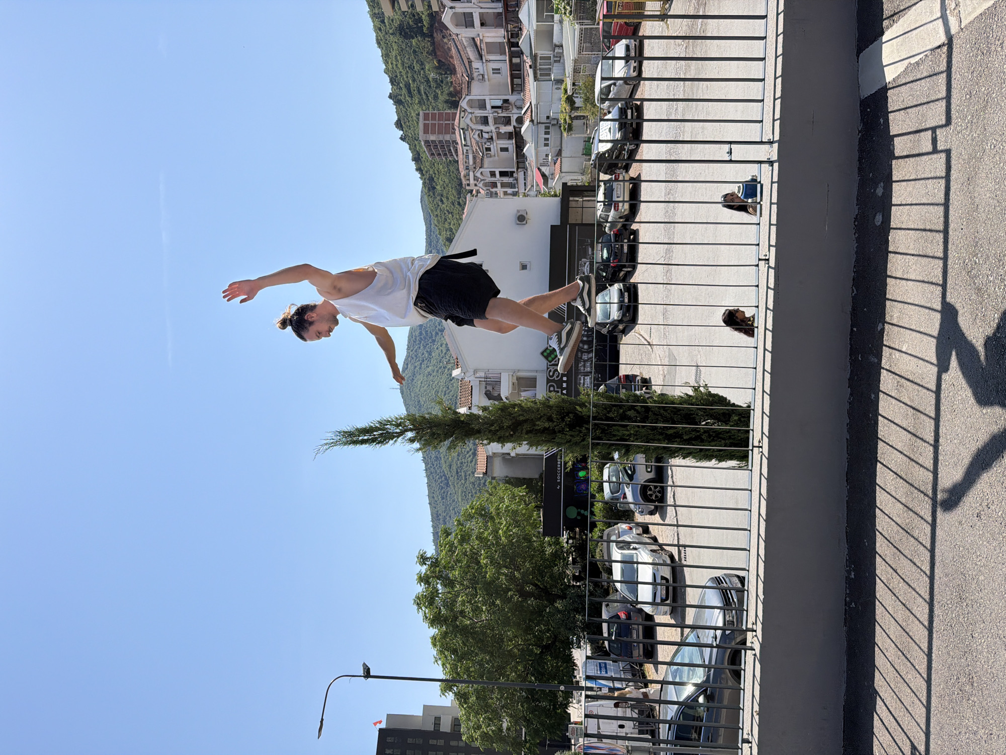 A vertically oriented outdoor shot features a man mid-air, seemingly leaping over a balcony rail. The man has a fair complexion, dark hair pulled back in a bun, and is wearing a white tank top and black shorts. He is turned towards the left, with arms outstretched.
The balcony has a metal railing with closely spaced vertical bars. Beyond the railing, the scene depicts a parking lot with multiple vehicles. The background is dominated by a hillside dotted with buildings, indicating an urban or suburban location. The sky is a clear blue, suggesting a sunny day. A tree is visible between the balcony and parking lot. A shadow, presumably of the photographer, is cast on the right edge of the image.
Based on the architecture and landscape, the photo appears to be taken in Budva, Montenegro.
