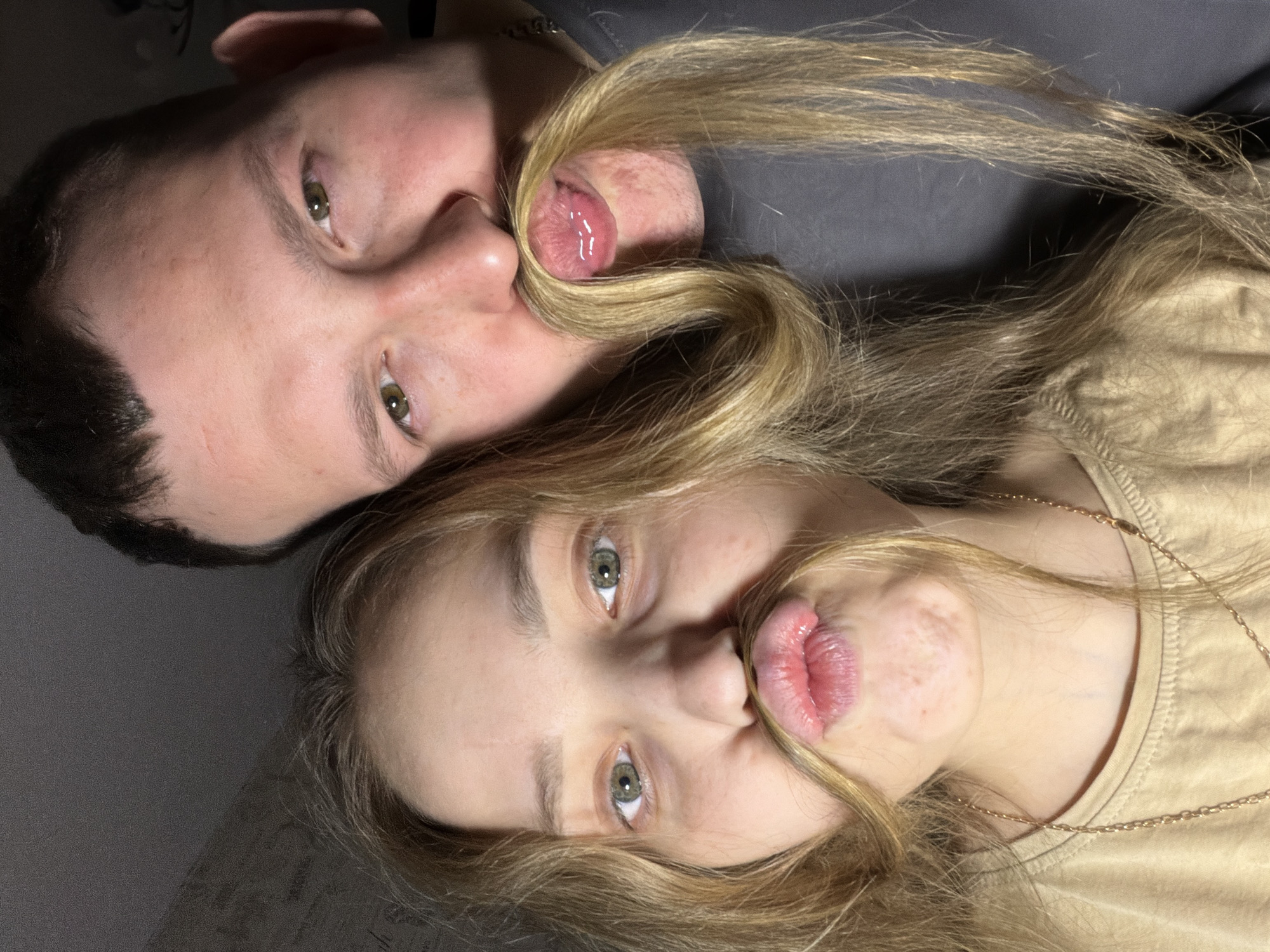 A couple is lying down, their faces close together. The man, on the left, has dark hair and green eyes. The woman, on the right, has blonde hair and green eyes. They are each holding strands of the woman's hair in their mouths, imitating moustaches. The man is wearing a dark grey shirt, and the woman is wearing a light brown shirt. There is a gold necklace visible around the woman's neck. The background is a light grey surface. It is impossible to confirm the location is Chaikovsky, Russia.