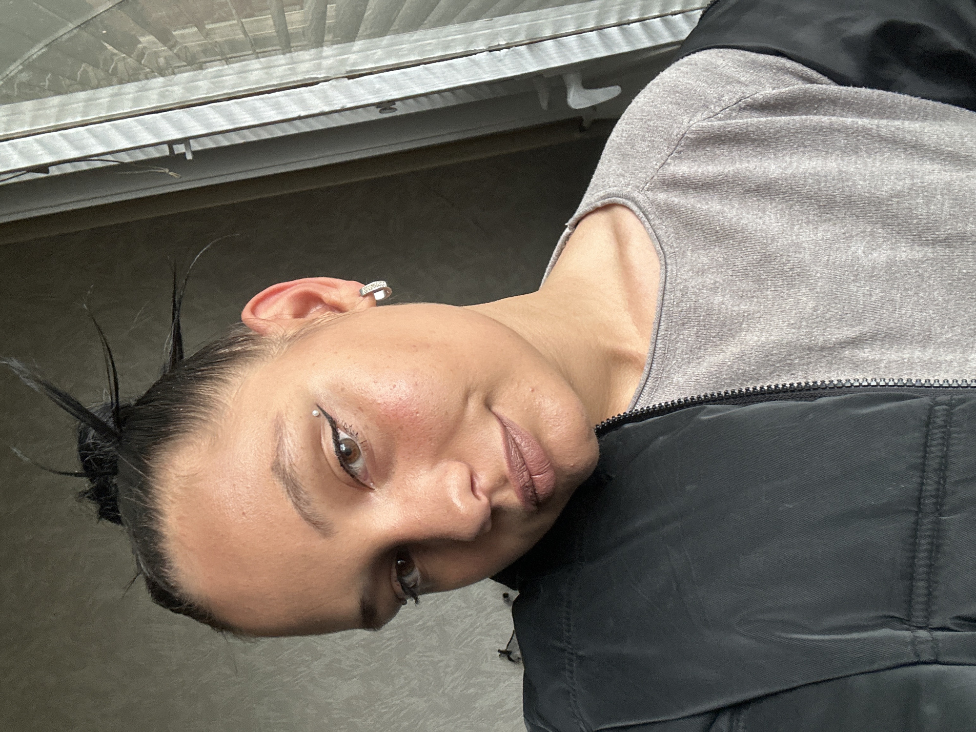 A woman with dark hair pulled back smiles slightly at the camera. She has a silver stud in her eyebrow and a geometric earring. She wears a gray, long-sleeved shirt and a black jacket. The background appears to be a window with a white frame. It is not possible to ascertain if this image was taken in Sofia, Bulgaria.