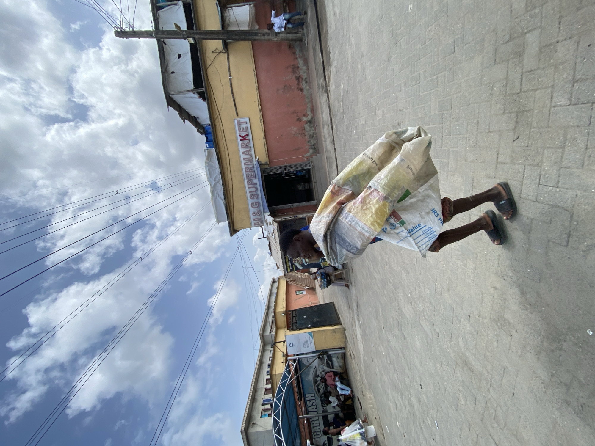 An urban street scene in Ado, Nigeria, is captured on a bright, partly cloudy day. The image shows a paved sidewalk and a street flanked by commercial buildings.
In the foreground, a person is walking, seen from the waist down. They wear dark sandals and are almost entirely covered by a large, light-colored, possibly woven plastic sack, which bears various texts and graphics, including "IRS PR", "Value", and what appears to be Tamil script (e.g., "அரிசி" for rice).
To the right, a two-story building features a prominent "M&G SUPERMARKET" sign in white letters on a blue background, above its entrance. The building has a yellow upper facade and a reddish lower section. On an upper-floor balcony of this building, a man in a white shirt is visible. Further down the street, another business with a "CAR WASH" sign is seen under a metal-framed shelter, where several people are gathered.
Numerous black electrical wires crisscross the sky, supported by a wooden utility pole. The sky is bright blue with scattered white clouds, indicating daytime, likely late morning or early afternoon. The overall setting depicts a busy commercial street environment.