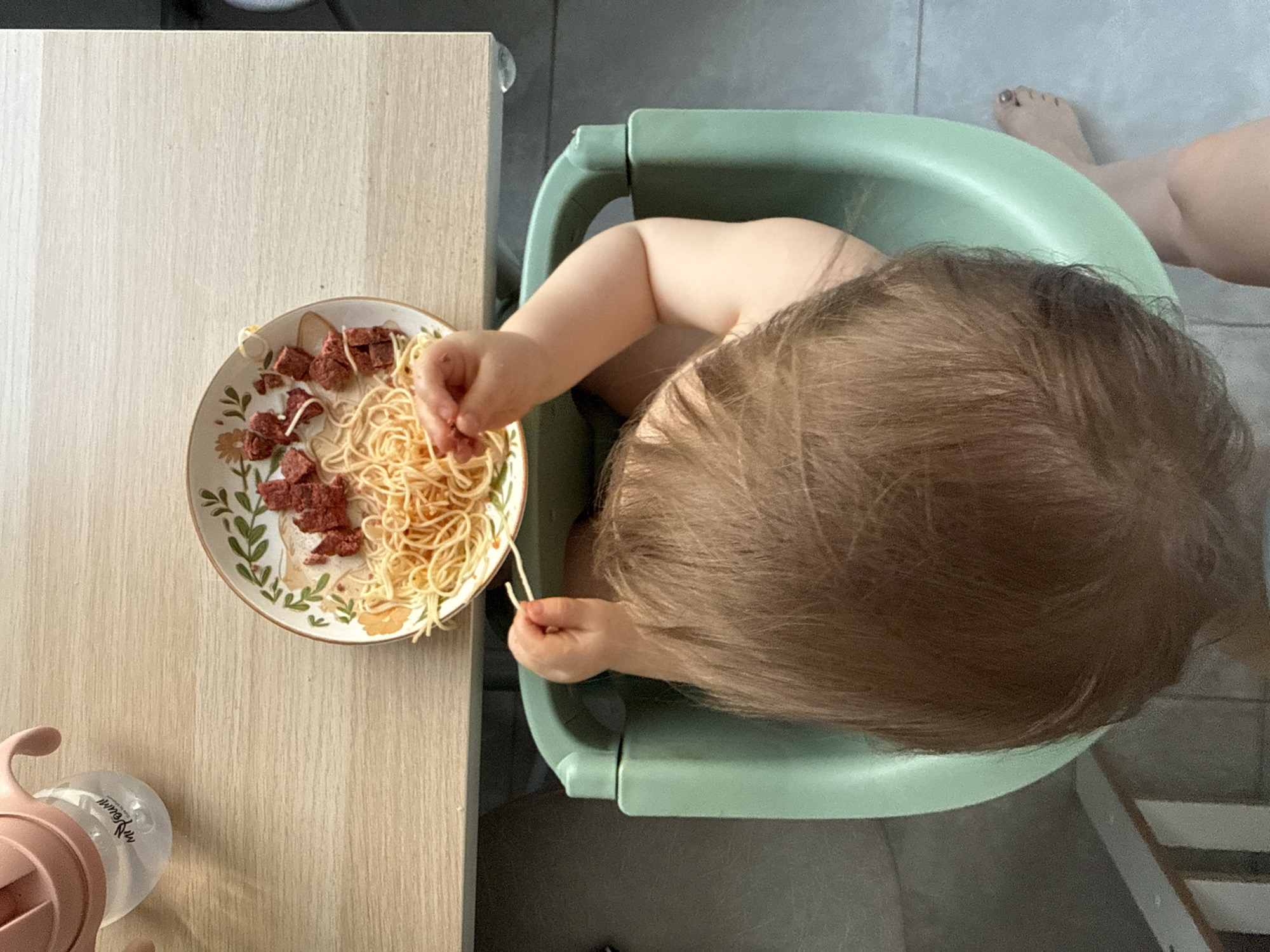 A top-down shot shows a baby sitting in a green high chair at a wooden table with a plate of spaghetti and diced meat. The baby is reaching for the spaghetti with its hands. A sippy cup is on the table next to the plate. A person's foot is visible in the top right of the frame. The location is not Moscow, Russia.