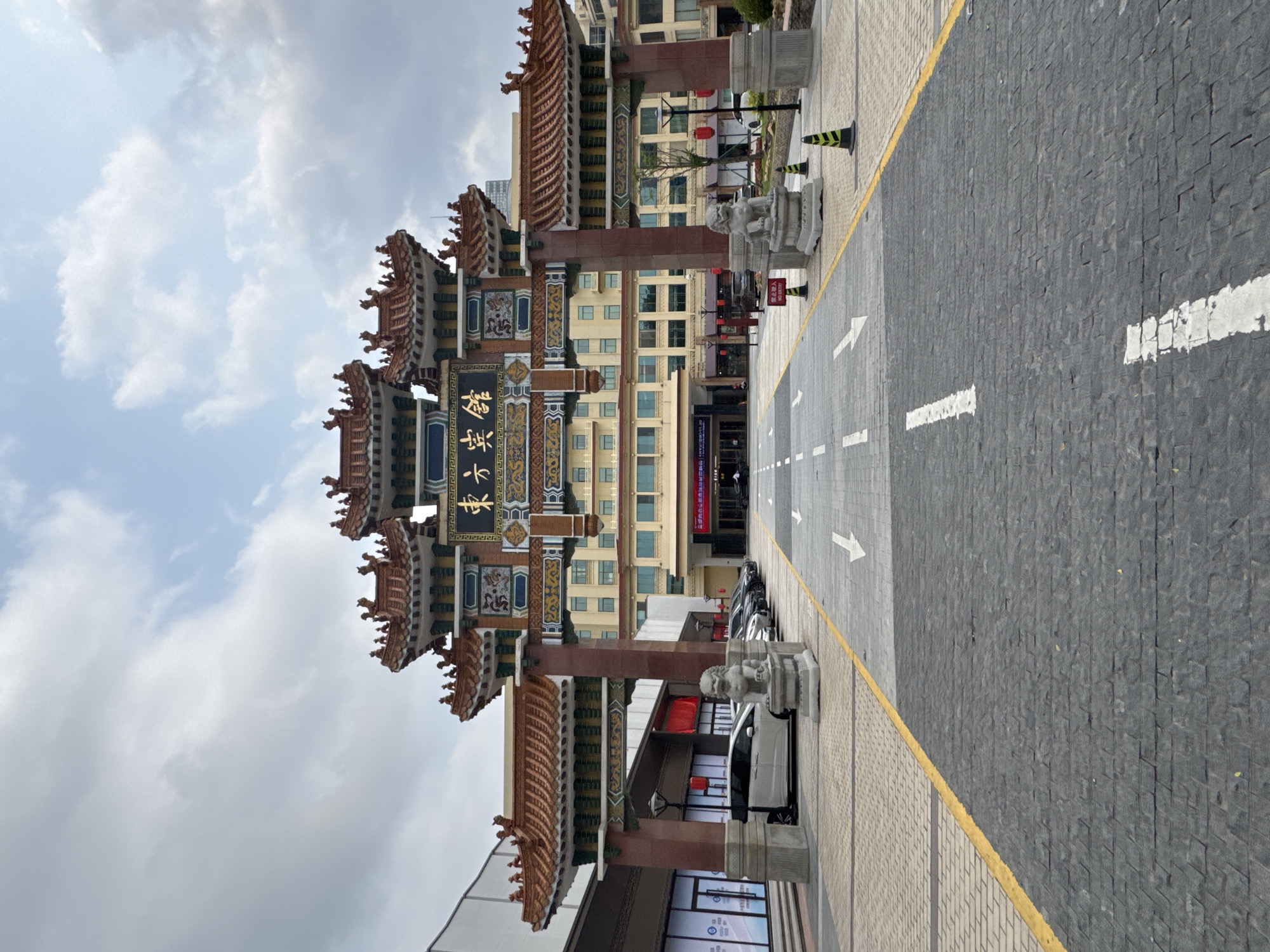 The image shows a street view in Yuexiu District, China, featuring a traditional Chinese Paifang gate. The gate is ornately decorated with multi-tiered roofs, intricate carvings, and Chinese script. It stands at the entrance to a street, likely a pedestrian zone. The street is paved with grey bricks and marked with white directional arrows. The sides of the street are lined with modern buildings. Parked cars are visible. The sky is overcast with white and grey clouds.