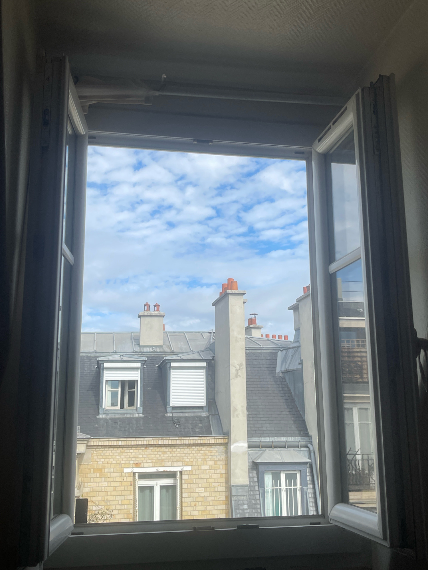 The image shows an interior view looking out of an open window at Parisian rooftops. The window frame is white and appears to be made of uPVC. The sky is blue with scattered white clouds. The rooftops are predominantly gray slate with visible chimneys, some topped with orange caps. The buildings are a mix of light beige brick and darker gray facades. Several windows with roll-down shutters are visible. The overall scene depicts a typical urban residential landscape in Paris, France. There are no people or animals visible in the image. The lighting suggests it is daytime, likely late morning or early afternoon.