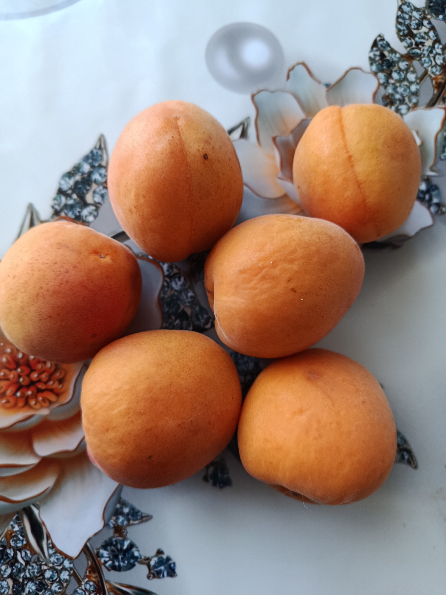 A close-up, overhead shot displays six apricots arranged on a light-colored surface adorned with a floral pattern. The apricots are primarily a warm orange hue, with subtle variations in color, some exhibiting a slight blush of red or yellow. They are positioned in a somewhat scattered, organic arrangement, with a few clustered together and others spaced further apart. The surface beneath the apricots features a design that appears to depict stylized flowers and possibly some decorative embellishments, such as what might be rhinestones or glitter, adding a touch of sparkle to the scene. The lighting suggests an indoor environment, likely natural light from a window, casting soft shadows that highlight the rounded forms of the fruit. The image does not contain any people or indicate a specific location within the city of Svietlahorsk, Belarus.