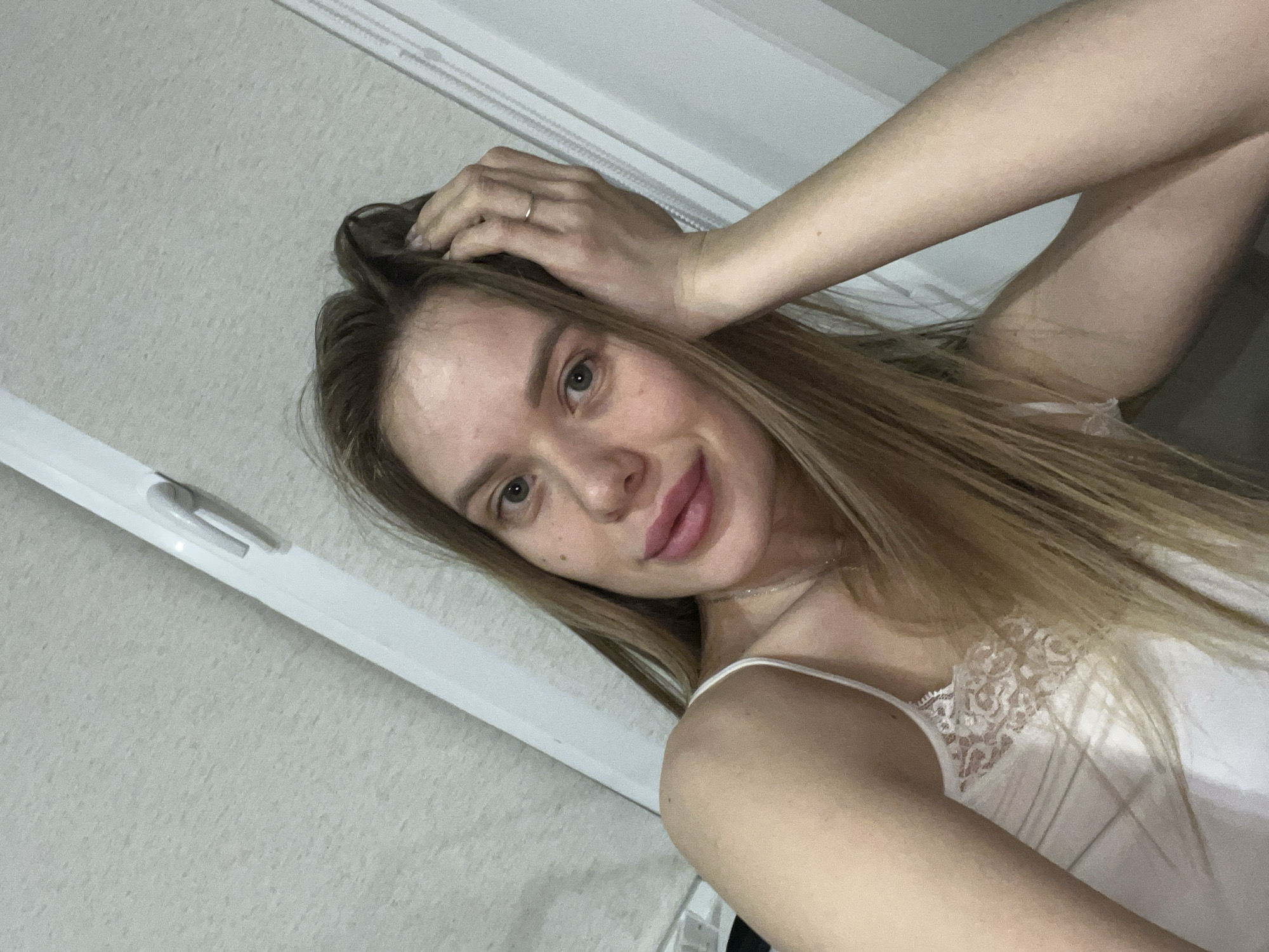 A light-skinned woman with long blonde hair poses for a selfie indoors. She has her hand on her head and wears a white lace-trimmed tank top. The background shows a gray wall and a white window frame. There is no evidence to suggest the image was taken in Chaikovsky, Russia.