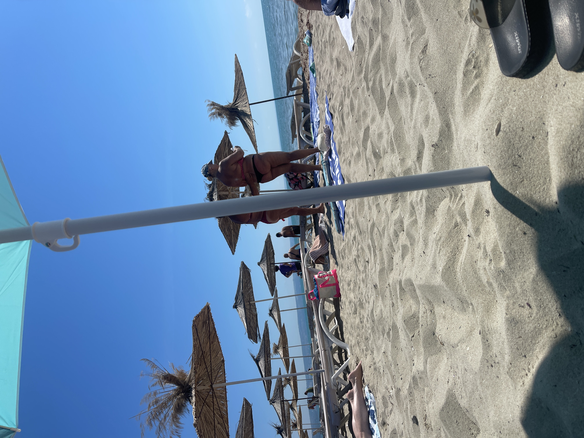A sunny day at the beach in Kranevo, Bulgaria, shows rows of thatched umbrellas and empty lounge chairs on a sandy shore. In the foreground, a turquoise beach umbrella pole casts a shadow. To the right, a pair of dark flip-flops rests on the sand. In the middle ground, two women in bikinis stand near a thatched umbrella. Further back, several people are seated on lounge chairs, and more umbrellas and chairs line the beach. The sea is calm with gentle waves under a clear blue sky.