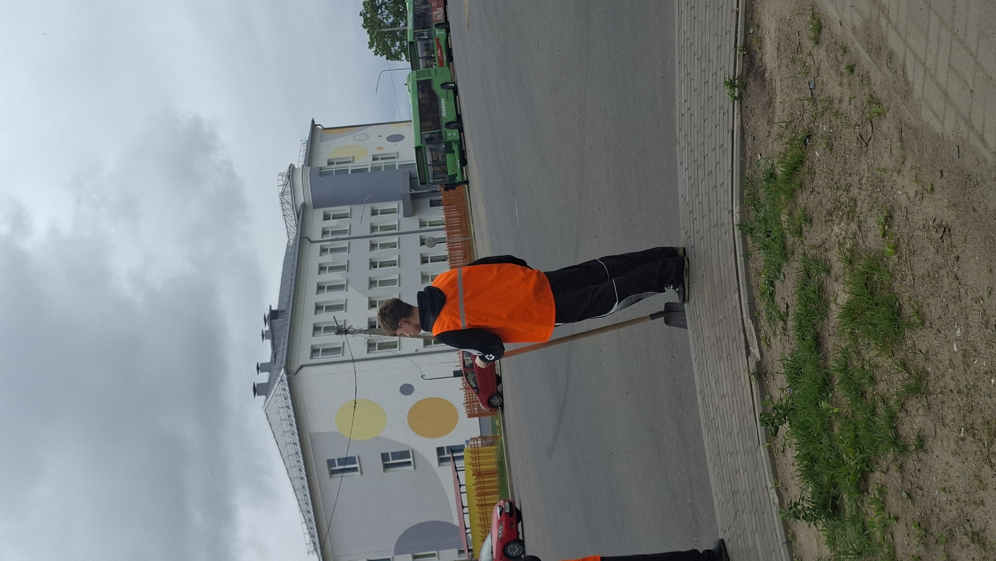 A male individual wearing a bright orange safety vest and black pants stands facing away from the camera, holding a shovel. He is positioned on a paved walkway beside a road. In the background, a large, multi-story building with a light-colored facade and circular decorative elements is visible. Several green buses are parked in a row on the road. The sky is overcast with dark clouds. The location is identified as Smalyavichy, Belarus.