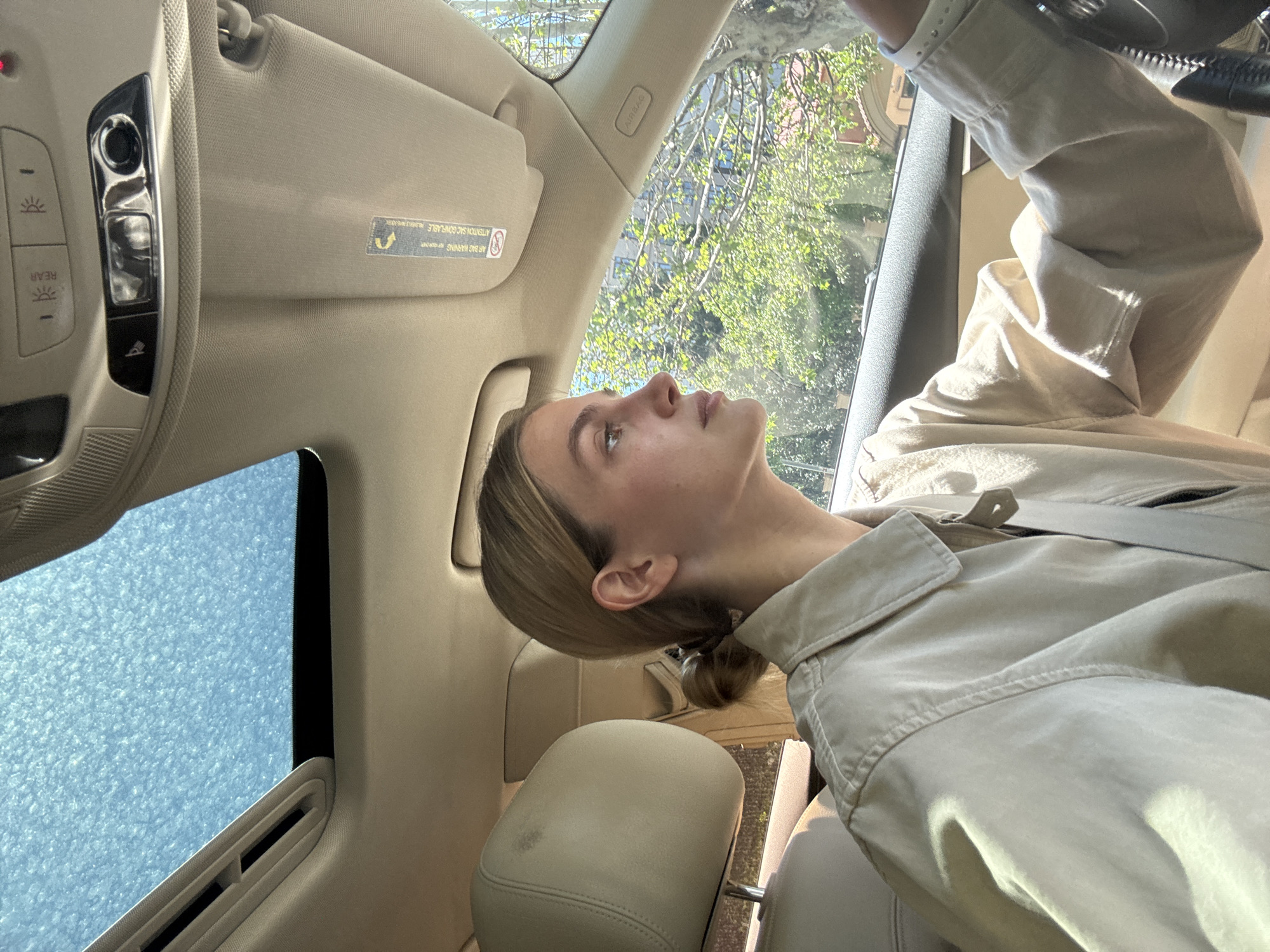 A fair-skinned woman with blonde hair pulled back in a low bun sits in the driver's seat of a car. She is looking upwards towards the sky. The car's interior is a light beige color. A sun roof is visible above her. Outside, trees with green leaves are visible through the car's windows. The city of Tbilisi, Georgia is not discernible in this image.