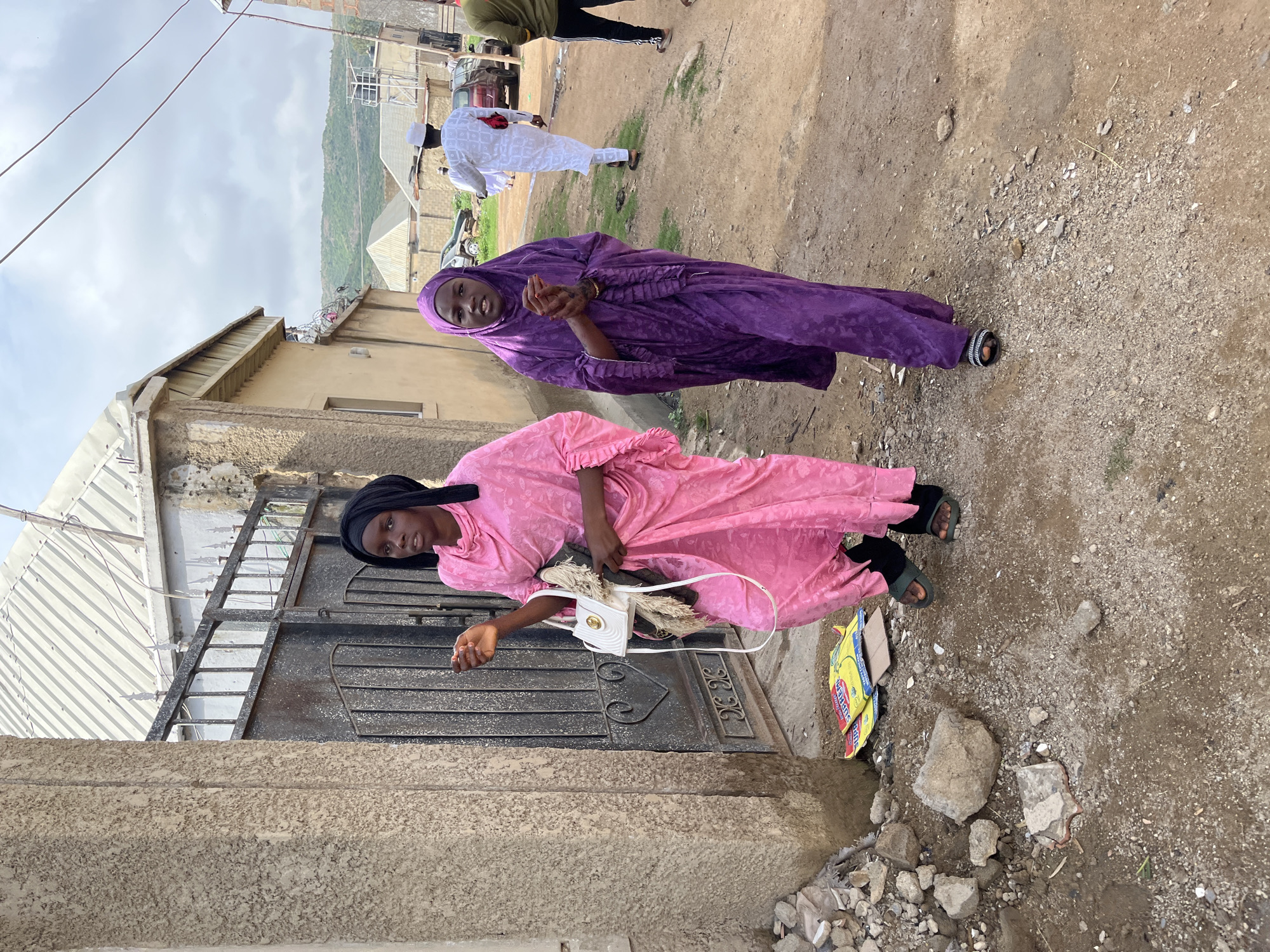 Two women in long dresses stand outside a building on a dirt road. One woman wears a pink dress with a black head covering and carries a white purse. The other woman wears a purple dress with a matching head covering. In the background, a man in white clothing walks along the road, and a car is visible further in the distance. The scene is in Jos, Nigeria, based on the environment, architecture, and clothing styles.