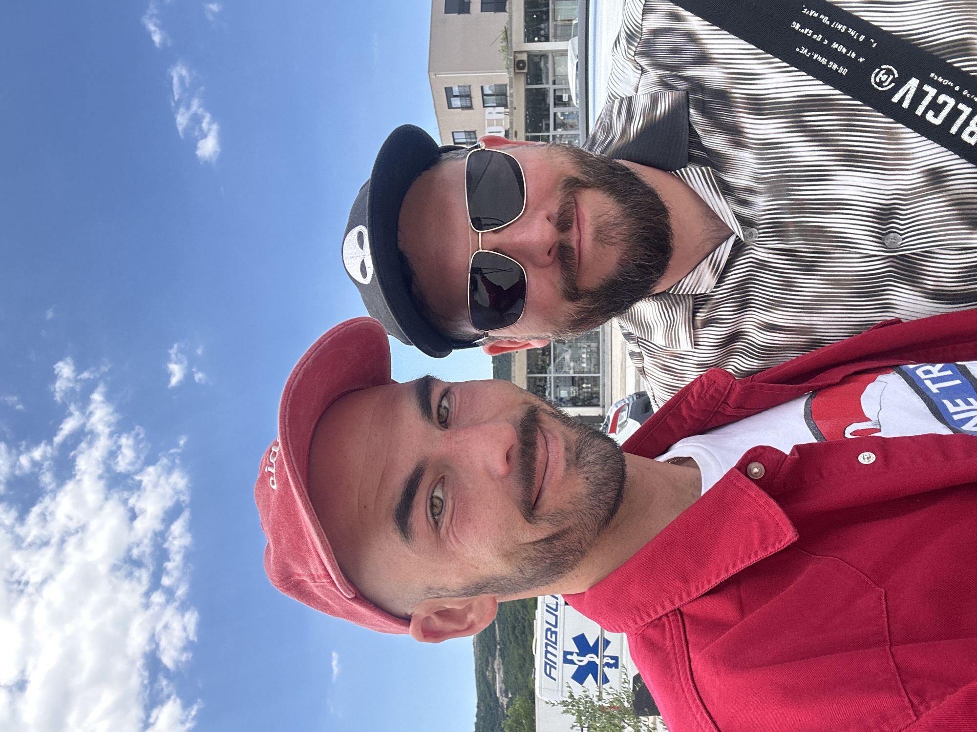 Two men are standing outside an "AMBULI" and "NE TR" building with a "BLCLV" shoulder strap. The man in the foreground is wearing a red cap and jacket with a t-shirt. The other man is wearing sunglasses and a patterned shirt. The image may be in Dub, Montenegro. The sky is blue with clouds.