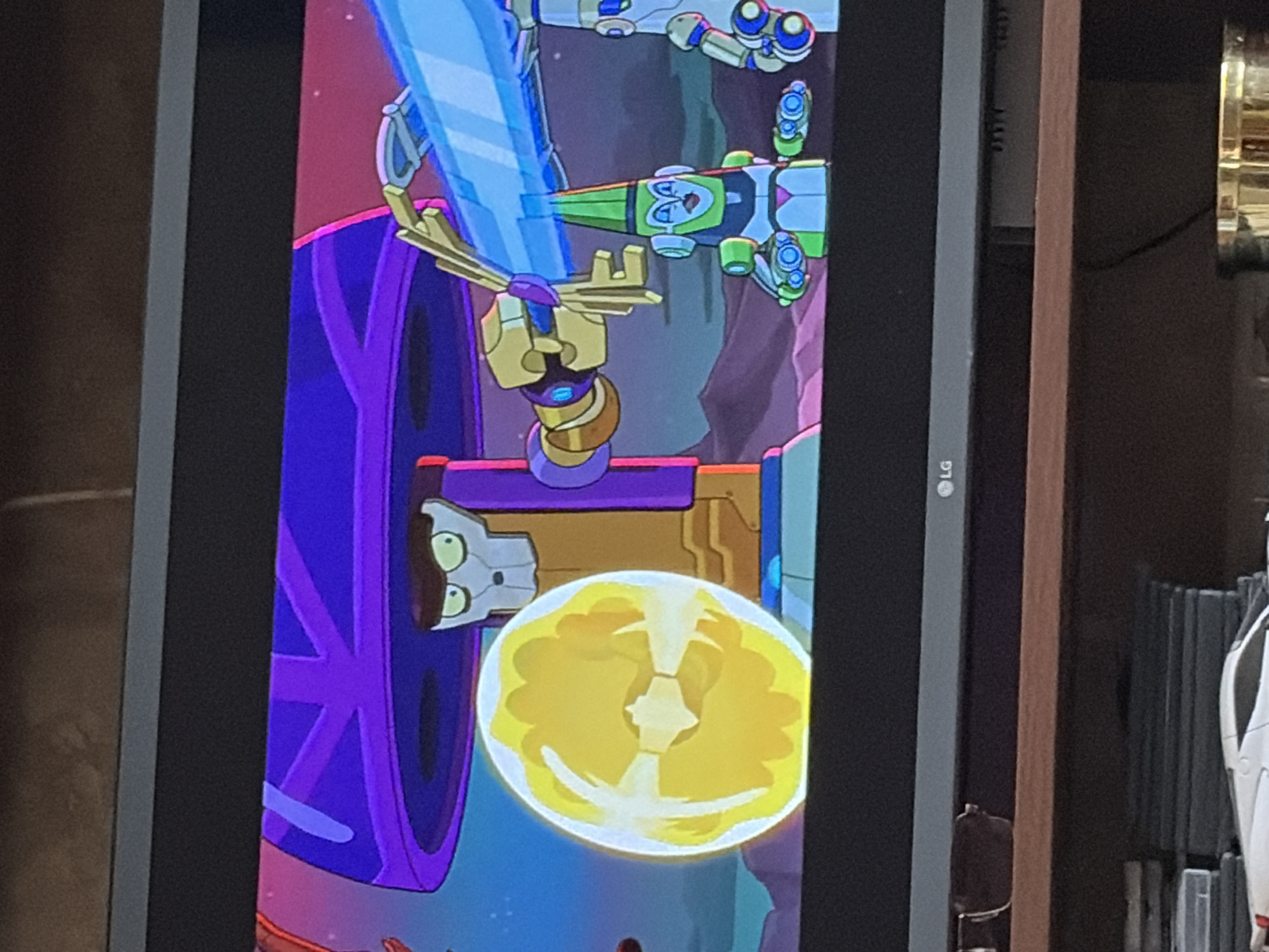 The image shows a scene from an animated cartoon, likely a science fiction show. In the foreground, a bright yellow, orb-like object with internal white patterns is centered. Behind this, a robot with a skull-like face and green eyes wearing glasses stands on a platform. A robotic arm extends from this robot, holding a large, blue, translucent blade or beam. To the right, a smaller, green and white robot with a cone-shaped hat is seated, flanked by two other robotic figures. The background depicts a purple structure, a pink sky, and a rocky, extraterrestrial landscape. The scene appears to be taking place in an artificial or alien environment. The bottom of the image shows a portion of a television screen with the LG logo visible. Below the television, a wooden shelf holds several objects, including a stack of what appear to be DVDs or game cases and the front of a white toy car. The lighting suggests the room is dimly lit.