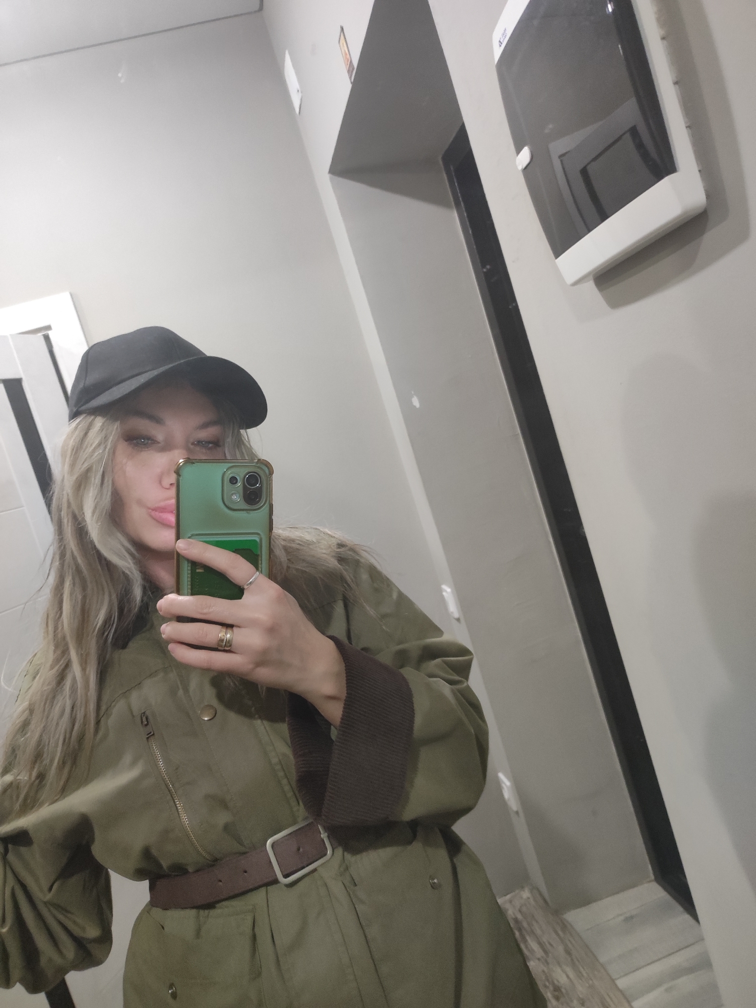 A woman is taking a selfie in a restroom. She is wearing a black baseball cap and a belted, olive-green jacket with brown cuffs. She is holding a green smartphone. The background shows a gray wall, a framed mirror, a black doorway, and a white dispenser mounted on the wall. The location is in Votkinsk, Russia.