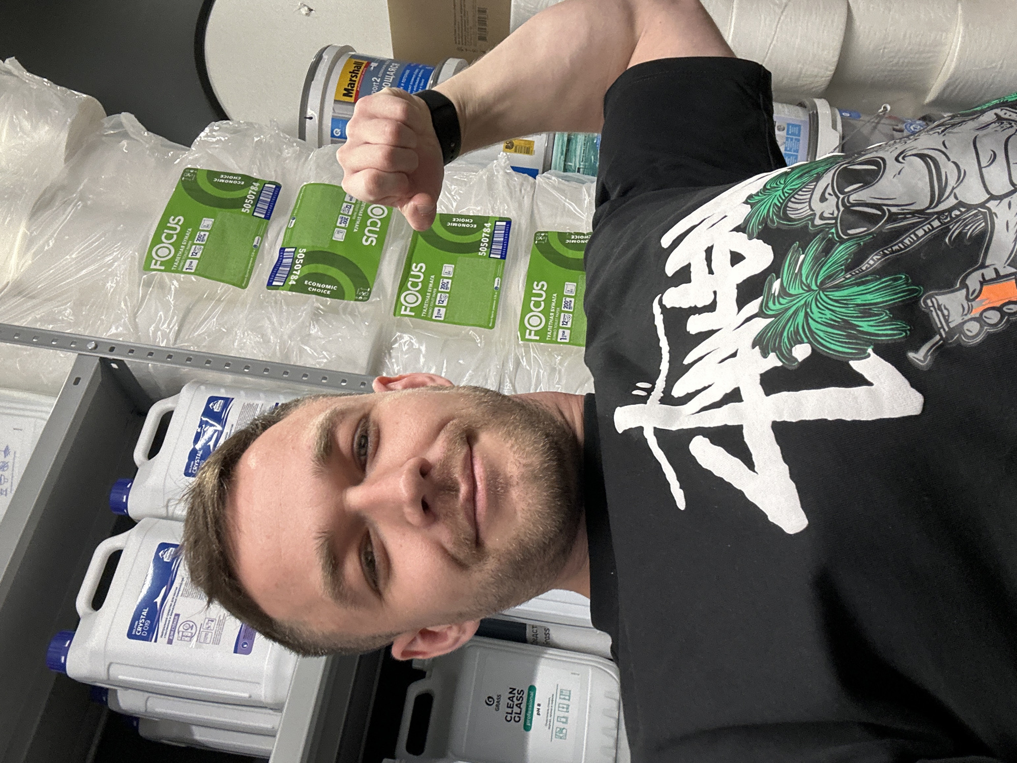 A man poses for a selfie in an indoor storage area, likely a warehouse or stockroom, in what appears to be Perm, Russia. He is fair-skinned, has short brown hair, and stubble. He is wearing a black t-shirt with a large white graphic design. The man is in the foreground, with his upper body and face clearly visible. His left arm is raised in a fist.
Behind him, a shelf unit is visible, stocked with various items. On the left side of the shelf, there are multiple white containers of "CRYSTAL" and "CLEAN GLASS" branded cleaning products, stacked on top of each other. Above the shelf, there are packages of "FOCUS" branded toilet paper. Rolls of what appear to be industrial paper towels or cleaning wipes are stacked on the top shelf along with a can of "Marshall" branded paint.
The "FOCUS" toilet paper packaging indicates they are "ECONOMIC CHOICE," and the text on the "CRYSTAL" cleaning product is in Russian. The city is inferred by the brands of products visible.