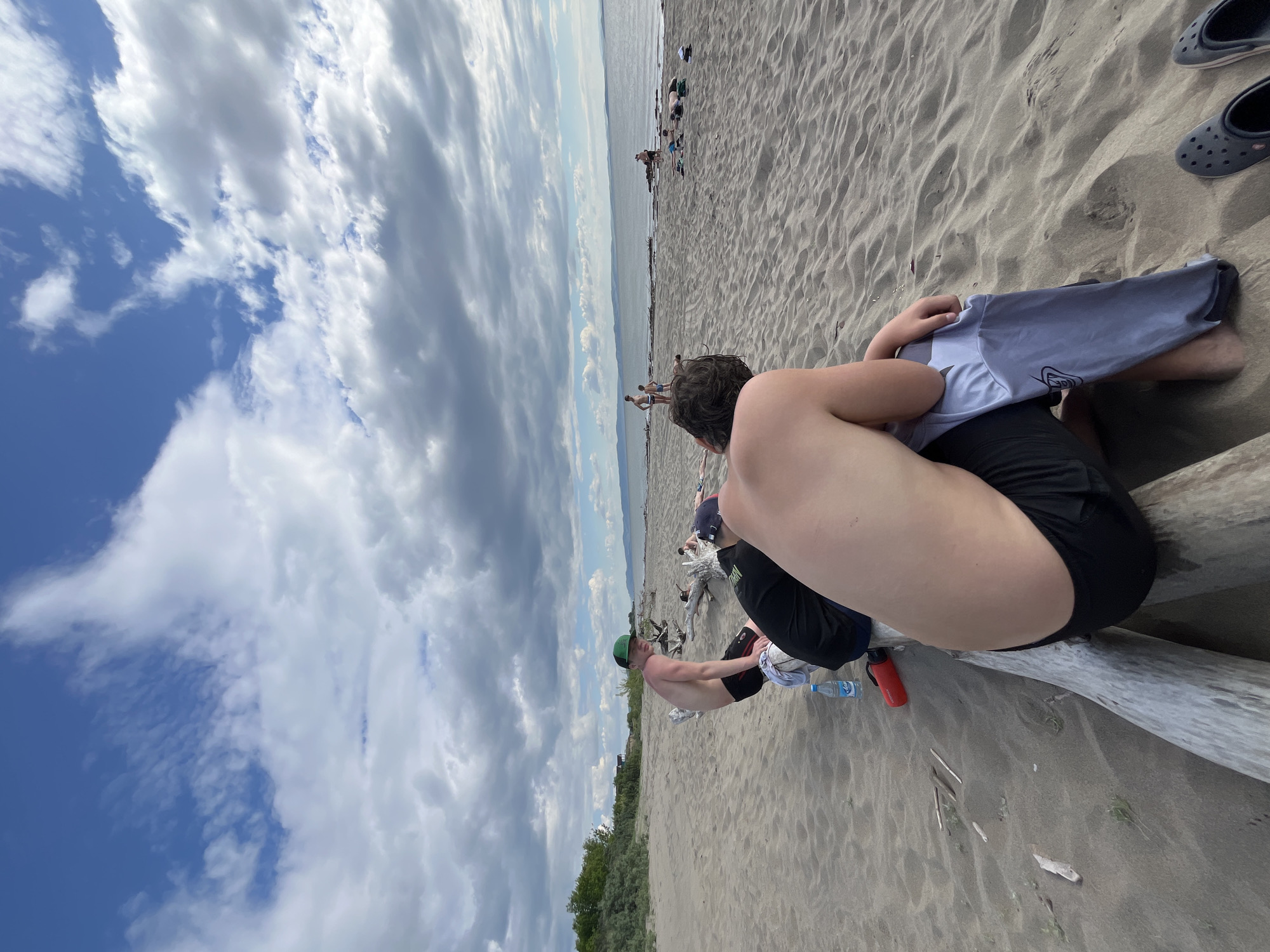 The image depicts a beach scene with several people in the foreground and background. In the foreground, a young man with dark curly hair is sitting with his back to the camera, wearing black shorts and holding a light gray t-shirt. To his left, another young man with fair skin and dark hair is also seated on a log, wearing black shorts and a black t-shirt. He is wearing a green baseball cap. A blue water bottle and a red thermos are visible on the sand near them.
In the mid-ground, a group of people are visible near the water's edge. Some are standing, and others appear to be lying down on the sand. The beach is sandy, with driftwood logs scattered around. The background shows a body of water, possibly a lake or sea, with hills or mountains in the distance under a cloudy sky. The overall environment appears to be a natural, outdoor setting, likely a beach. The specific location in Krasnoturansk, Russia, is not visually identifiable from the image content alone.