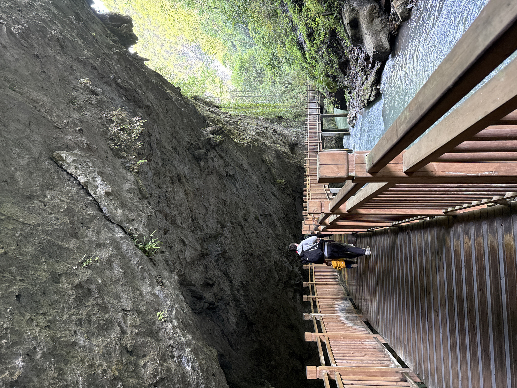 The image shows a person walking on a wooden walkway suspended along a cliff face in Zhangjiajie, China. The walkway runs alongside a river or stream below. To the left of the walkway is a steep, rocky cliff face with sparse vegetation. Above the cliff and river, trees and foliage are visible. The person is wearing a black jacket, a light-colored hoodie, dark pants, and white sneakers. They have a yellow bag. The walkway is constructed of wooden planks and has wooden railings. It appears to be a tourist attraction.