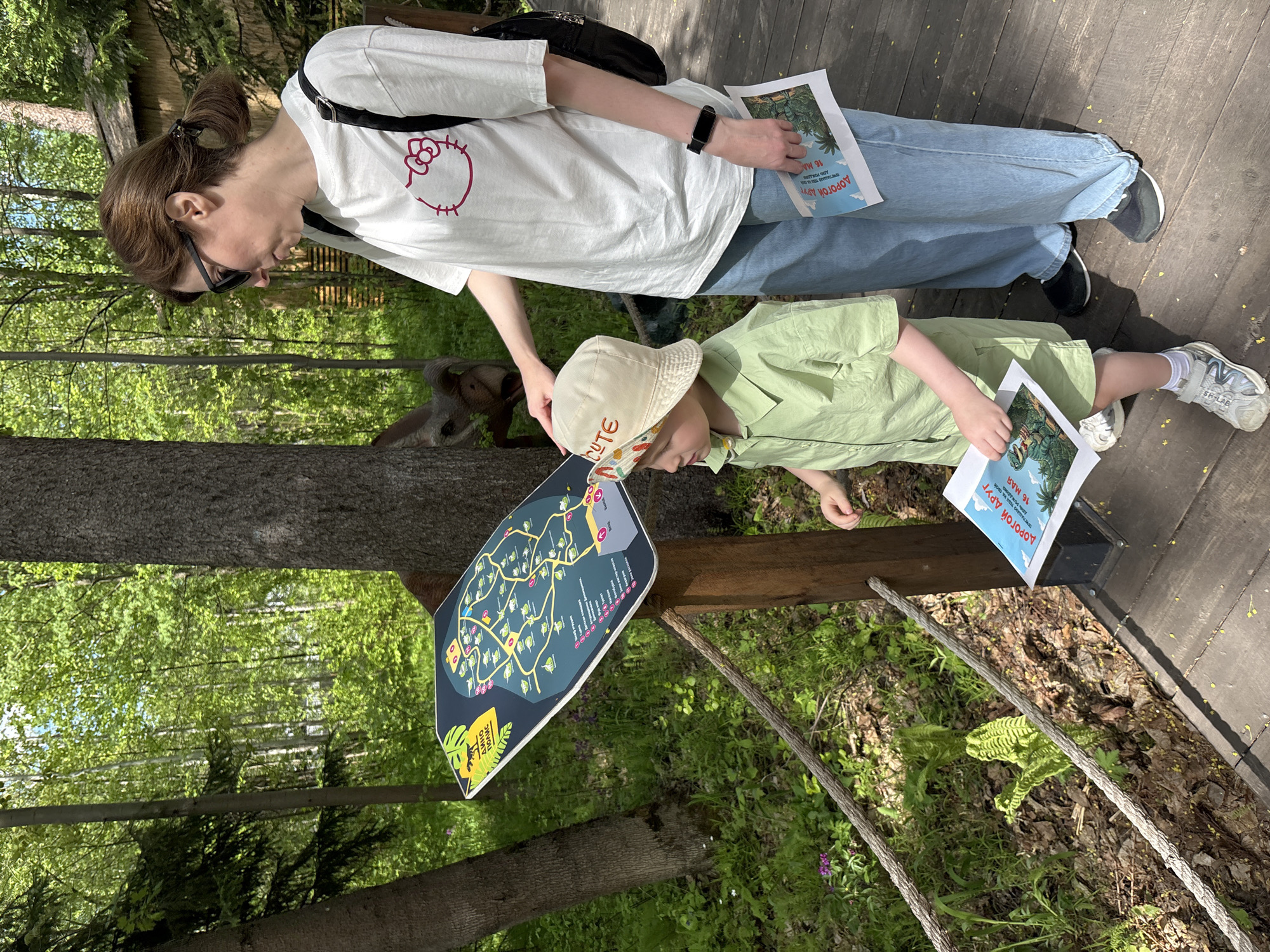 The image shows an adult and child viewing a park map in what appears to be a wooded area. The adult, a fair-skinned woman, is wearing a white t-shirt with a Hello Kitty logo, blue jeans, and black sneakers. She has her brown hair pulled back in a ponytail and is wearing sunglasses. She holds a brochure in her hand and a black bag is slung over her shoulder.
The child, presumably a young boy, is wearing a light green shirt and khaki pants, along with white and gray sneakers. He wears a light beige bucket hat with the word "CUTE" on it. He is also holding a brochure similar to the one held by the adult. Both the adult and child are looking at a park map showing different dinosaur exhibits, labelled with the word "Dinopolis".
The background is composed of trees, wooden planks suggesting a walkway, and forest floor. The map is mounted on a wooden railing with rope strung across it. The park is likely located in Izhevsk, Russia based on some text on the brochure, and the general look of the landscape.