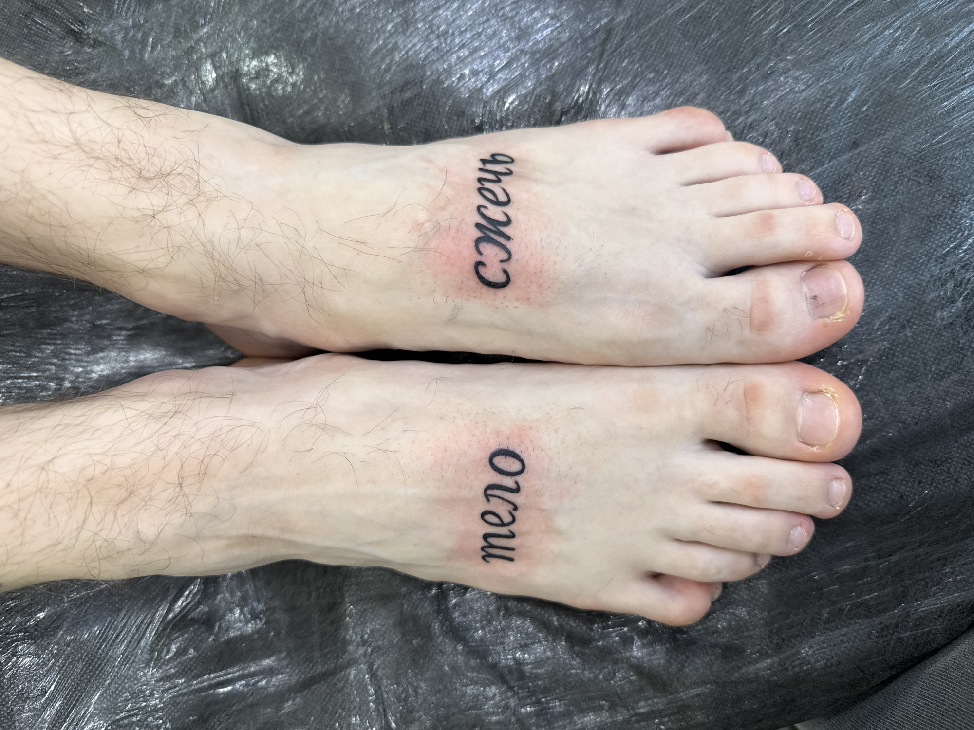 This close-up, overhead photograph displays the bare feet and lower shins of an individual, likely male, resting on a dark, crinkled surface, which appears to be a protective covering in a tattoo studio in Moscow, Russia. Both feet feature fresh, black ink tattoos, each oriented vertically and surrounded by noticeable redness and slight swelling, indicative of a very recent tattooing session.
On the right foot (viewer's left), the word "Сжечь" (Szhyech), meaning "Burn" in Russian, is tattooed in a flowing, script-like Cyrillic font. On the left foot (viewer's right), the word "тело" (telo), meaning "body" in Russian, is tattooed in a similar style. The skin on the shins and tops of the feet shows light-colored hair. The toenails are natural, unpolished, and some exhibit minor discoloration or unevenness. The overall image captures the immediate aftermath of new body art.