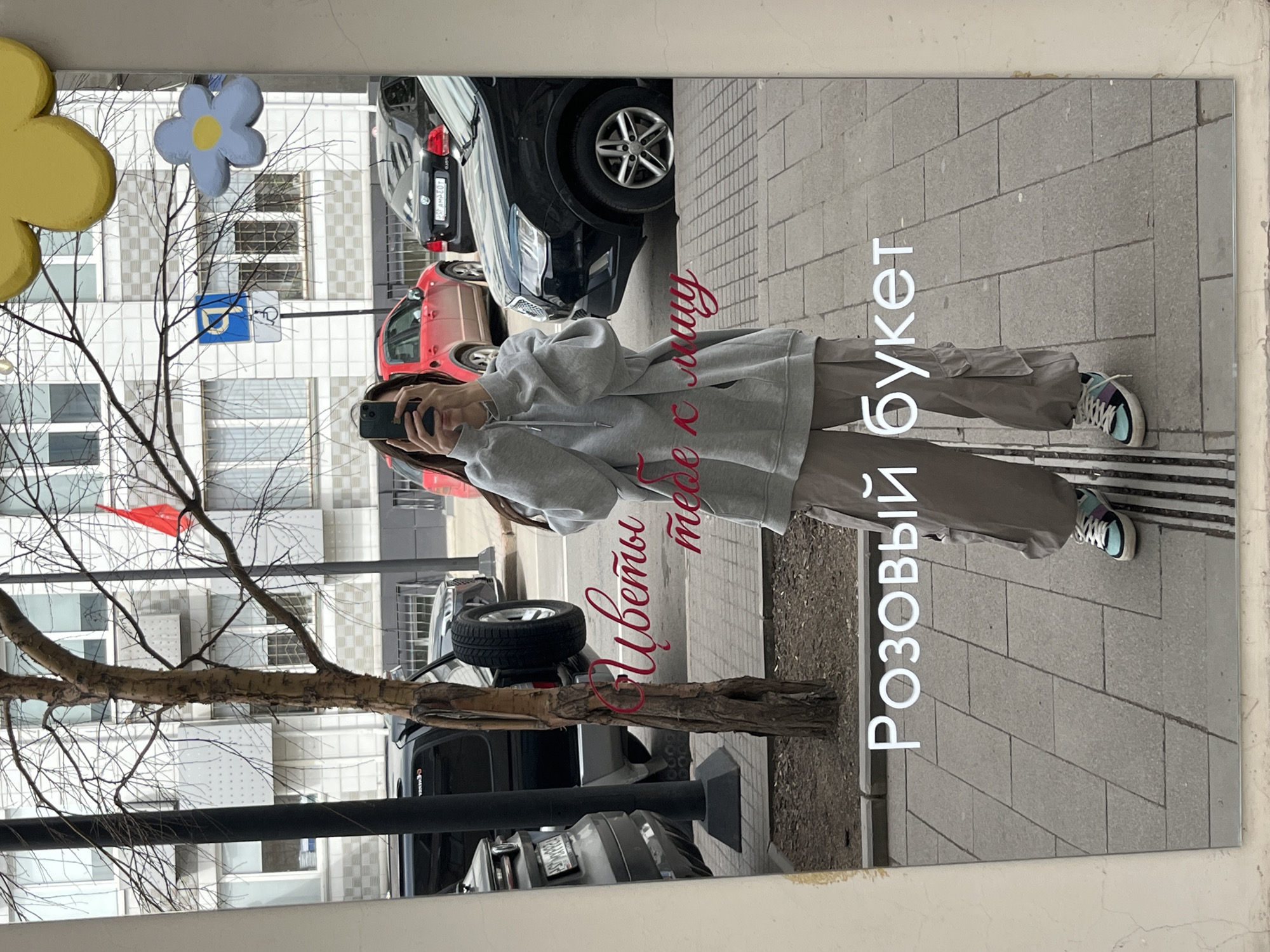 The image captures a reflection in a mirror, showing a woman taking a selfie. Behind her are several parked cars, and a building with multiple windows. A bare tree branch and some artificial floral decorations are also visible near the mirror. The reflection also contains Russian text: "Цветы тебе к лицу" and "Розовый букет", translating to "Flowers suit you" and "Pink bouquet". This photo was likely taken in Krasnoyarsk, Russia, based on the location of this shop.