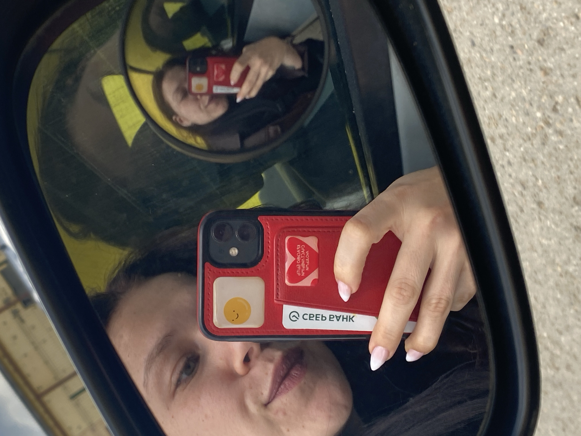 A close-up selfie is captured through a car's side mirror, prominently featuring a person's reflection holding a smartphone. The main mirror shows the person's face, with a neutral to slight smile, and their hand holding a red smartphone. Within this main reflection, a smaller, circular auxiliary mirror reflects the person's upper body and the phone again, creating a layered visual effect.
The smartphone is red with a black dual-camera module. Its red case features integrated card slots. One slot holds a white card with a yellow, smiling egg design. Another card, partially visible, is white with black text clearly reading "СБЕР БАНК" (Sberbank). Behind this, a red card shows reversed text, including "VOYAGER" and partially discernible Cyrillic words like "ПУТЕВОДИТЕЛЬ" (Guidebook) and "ЛУЧШИХ ВПЕЧАТЛЕНИЙ" (Best Impressions), suggesting a travel or loyalty program card from the Russian bank. The person's hand has manicured nails with a white French tip.
The setting appears to be outdoors during daylight hours, with a textured background resembling asphalt or a paved surface visible outside the car. The visible "Sberbank" text aligns with the location context of Chaikovsky, Russia.