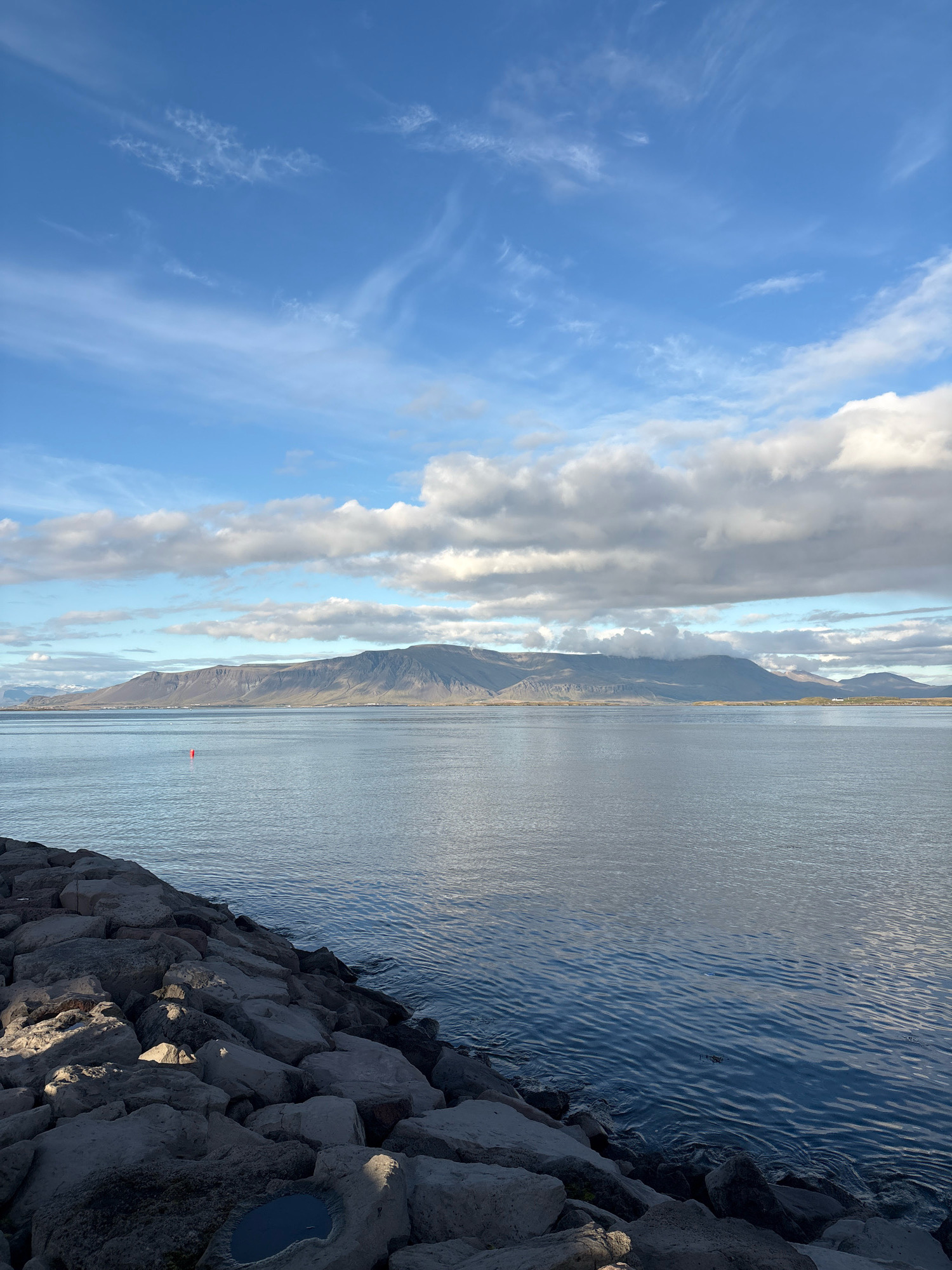 This image depicts a scenic view of the ocean with a mountainous backdrop, likely taken from a rocky shoreline. In the foreground, large, grey rocks form a breakwater, with a small pool of water visible among them. The ocean stretches out to the horizon, appearing calm with gentle ripples. A single red buoy floats in the water. In the distance, a long mountain range runs across the horizon, its peaks partially obscured by clouds. The sky above is a mix of blue and white, with wispy clouds scattered across it. This view is consistent with coastal areas of Reykjavik, Iceland, known for its proximity to dramatic natural landscapes. There are no people or animals visible in the image. The scene is static and appears to be captured during daylight hours.