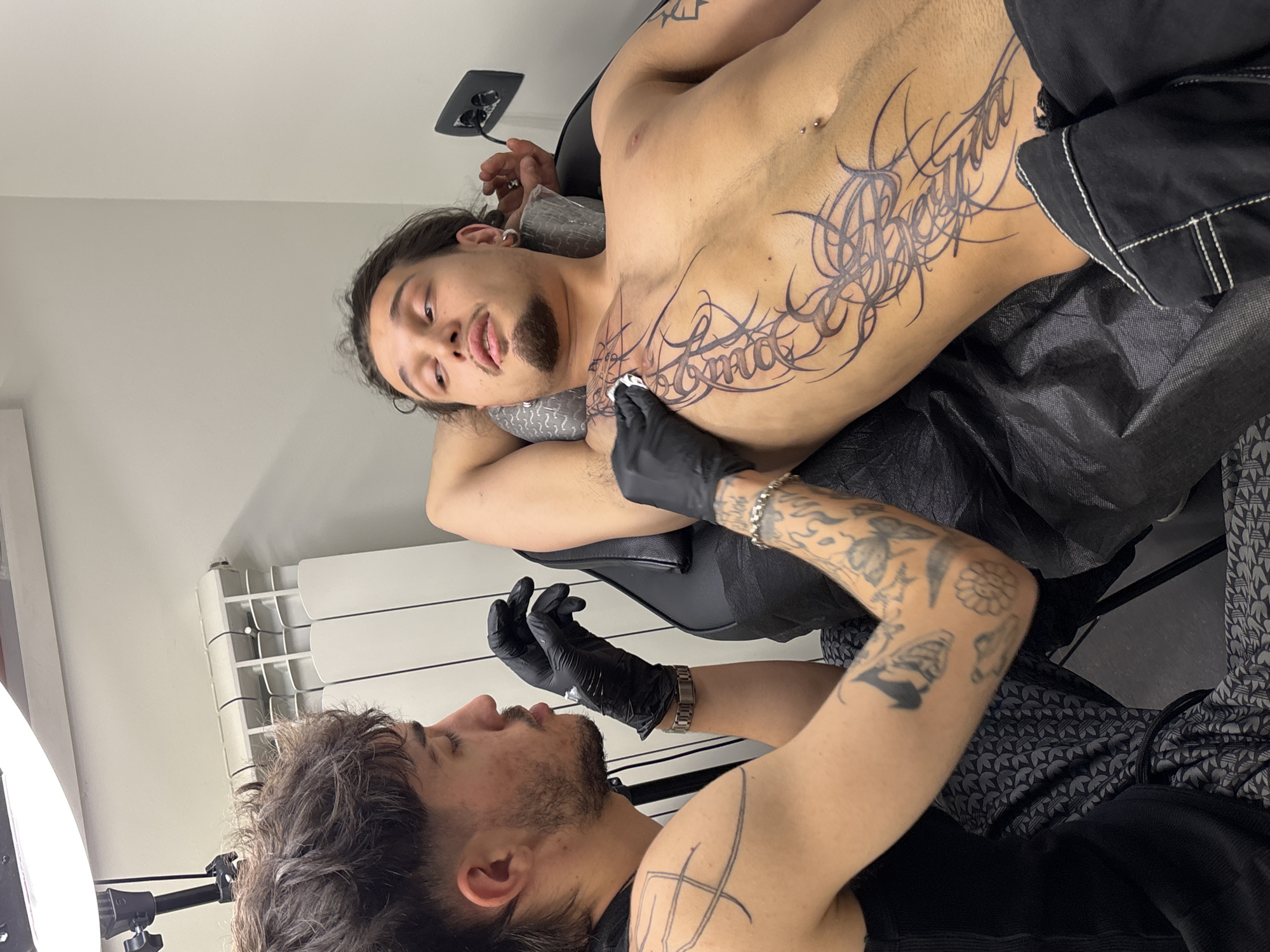 A tattoo artist, wearing black gloves, is in the process of tattooing a design onto the chest of a young man lying on a tattoo chair. The man has long dark hair and is looking up and to his left. The tattoo design is intricate and spans across his chest and upper abdomen. The environment appears to be a tattoo studio, with a radiator visible in the background and a bright light source overhead. The image does not provide any specific visual cues or landmarks that would allow for the identification of its location within the city of Sofia, Bulgaria.