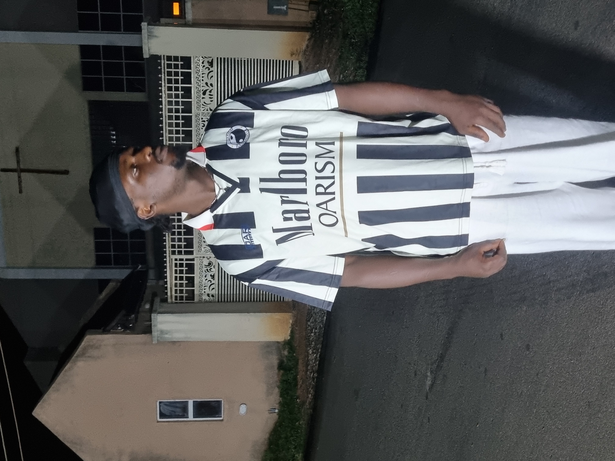 A man is standing on a paved surface in Jos, Nigeria. He is wearing a black head covering and a white and black striped soccer jersey with "Marlboro" and "OARISM" printed on it. He is also wearing white trousers. The background shows a building with a cross attached to its exterior. There is also a fence visible. The setting appears to be at night.