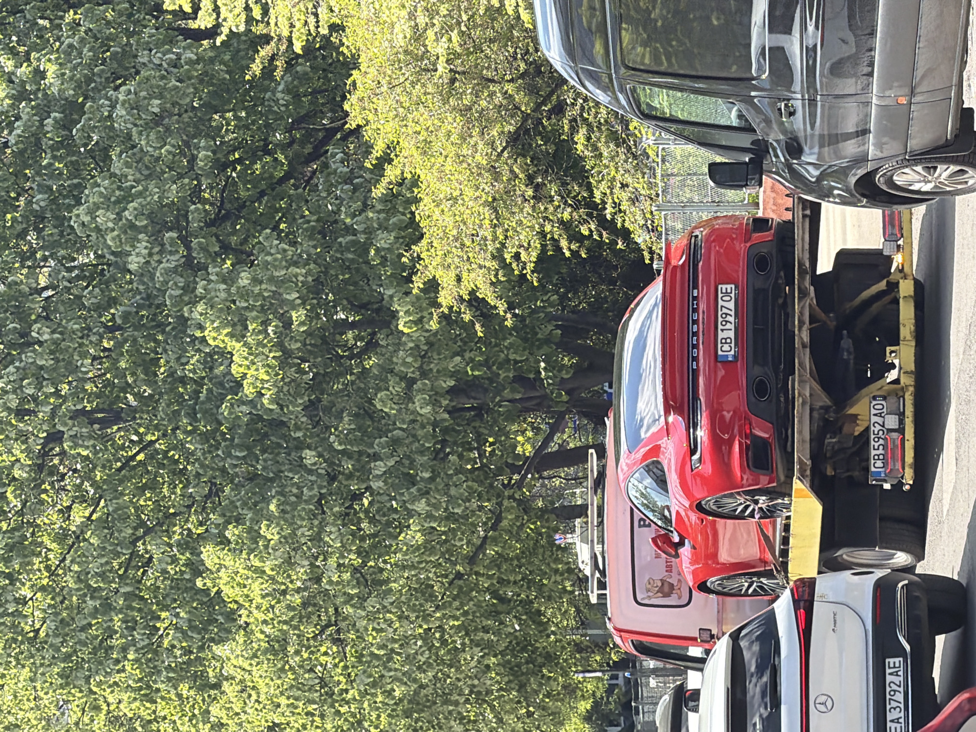 The image shows a red Porsche with license plate "CB 1997 OE" loaded on a yellow tow truck with license plate "CB 5952 AO." There's a black van to the right, and a white Mercedes with license plate "EA 3792 AE" partially visible at the bottom. The backdrop includes a large, leafy tree. The location appears to be a street in Sofia, Bulgaria, based on the license plates.