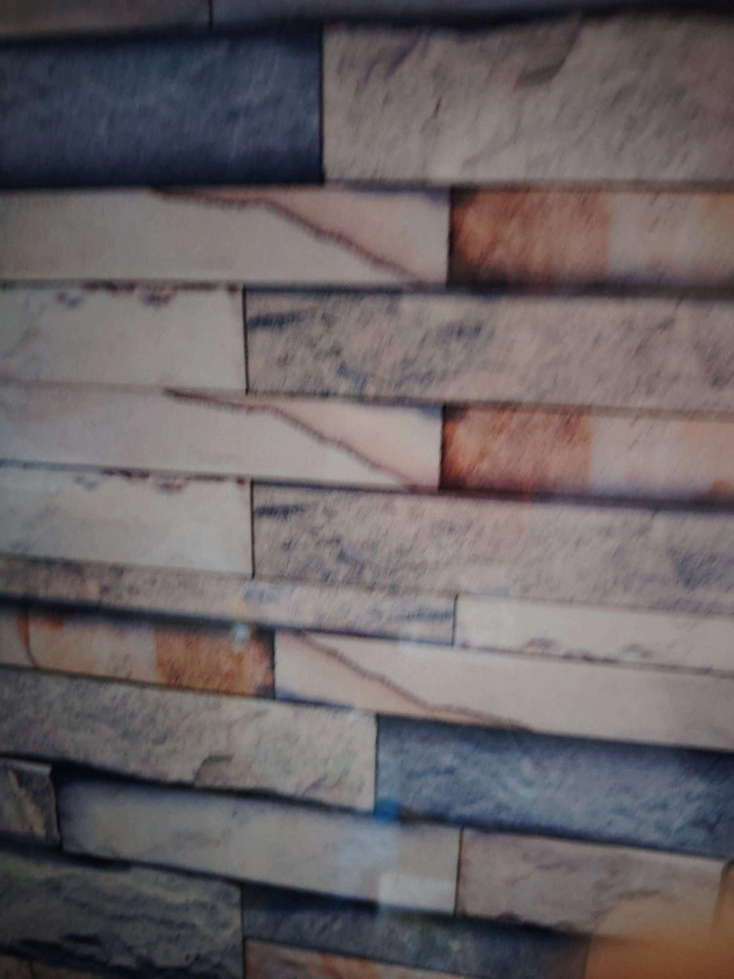 The image displays a close-up, horizontal, and slightly angled view of a stone veneer wall. The veneer consists of rectangular stones of varying sizes and colors, laid in a staggered pattern. The colors range from light beige and off-white to shades of gray, blue, and reddish-brown. Some stones have rough, textured surfaces with visible grain and minor imperfections. The lighting appears to be diffused, creating soft shadows and highlighting the textures of the stone. The image lacks any context of a specific location within the city of Huntingdonshire, United Kingdom, and contains no discernible people, objects, actions, or interactions.