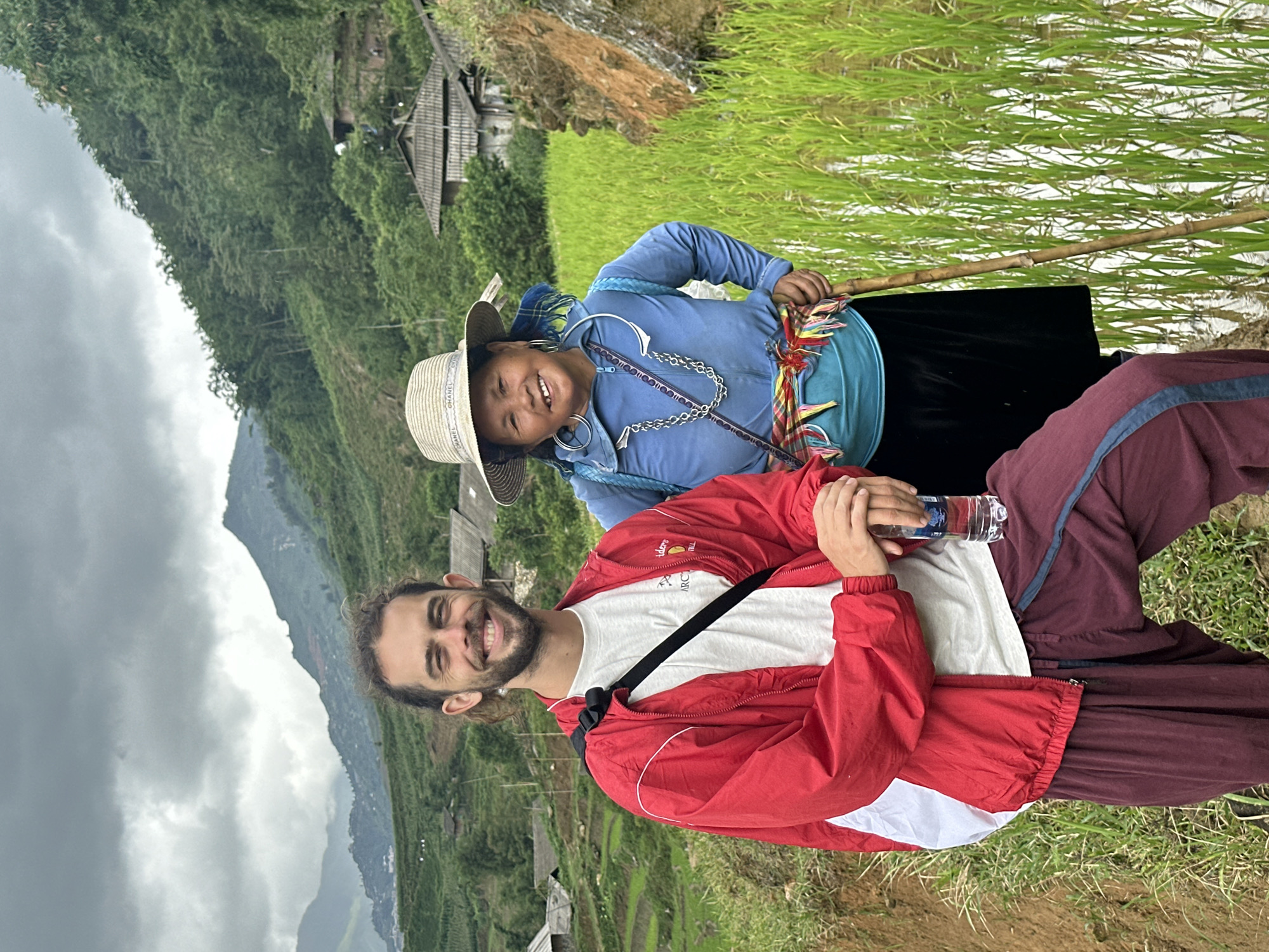 Two individuals are pictured in a mountainous region, likely within or near Mường Hoa, Vietnam, given the characteristic rice terraces visible in the background. The younger person, dressed in traditional blue clothing and a Chanel-branded straw hat, smiles towards the camera and holds a wooden stick. An adult male, wearing a red jacket, white t-shirt, and maroon pants, is also smiling while holding a water bottle. The setting includes vibrant green rice paddies and distant mountains under an overcast sky, suggesting a rural or agricultural area.