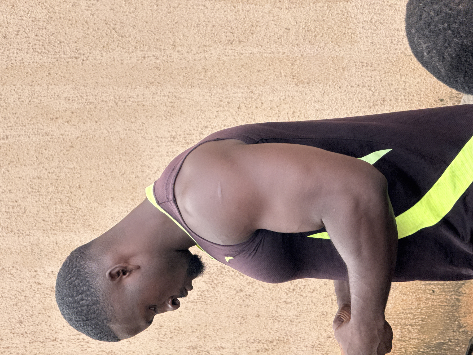 An African man is captured in a medium shot, looking down to his left. He is wearing a dark sleeveless athletic shirt with lime green accents along the sides and around the neck. The man has short, dark hair, a beard, and is facing a wall. The wall is textured and a light brown or beige color. His right arm is bent at the elbow, and his left hand is holding something small and dark in front of him, possibly a phone or wallet. His muscles are visible, suggesting he might be a bodybuilder or athlete. There is a faint scar on his right upper arm. The overall lighting is natural and suggests an outdoor setting. There is another dark, fuzzy object in the lower right corner of the image, possibly the back of someone's head. The background suggests the man is in Akwanga, Nigeria.