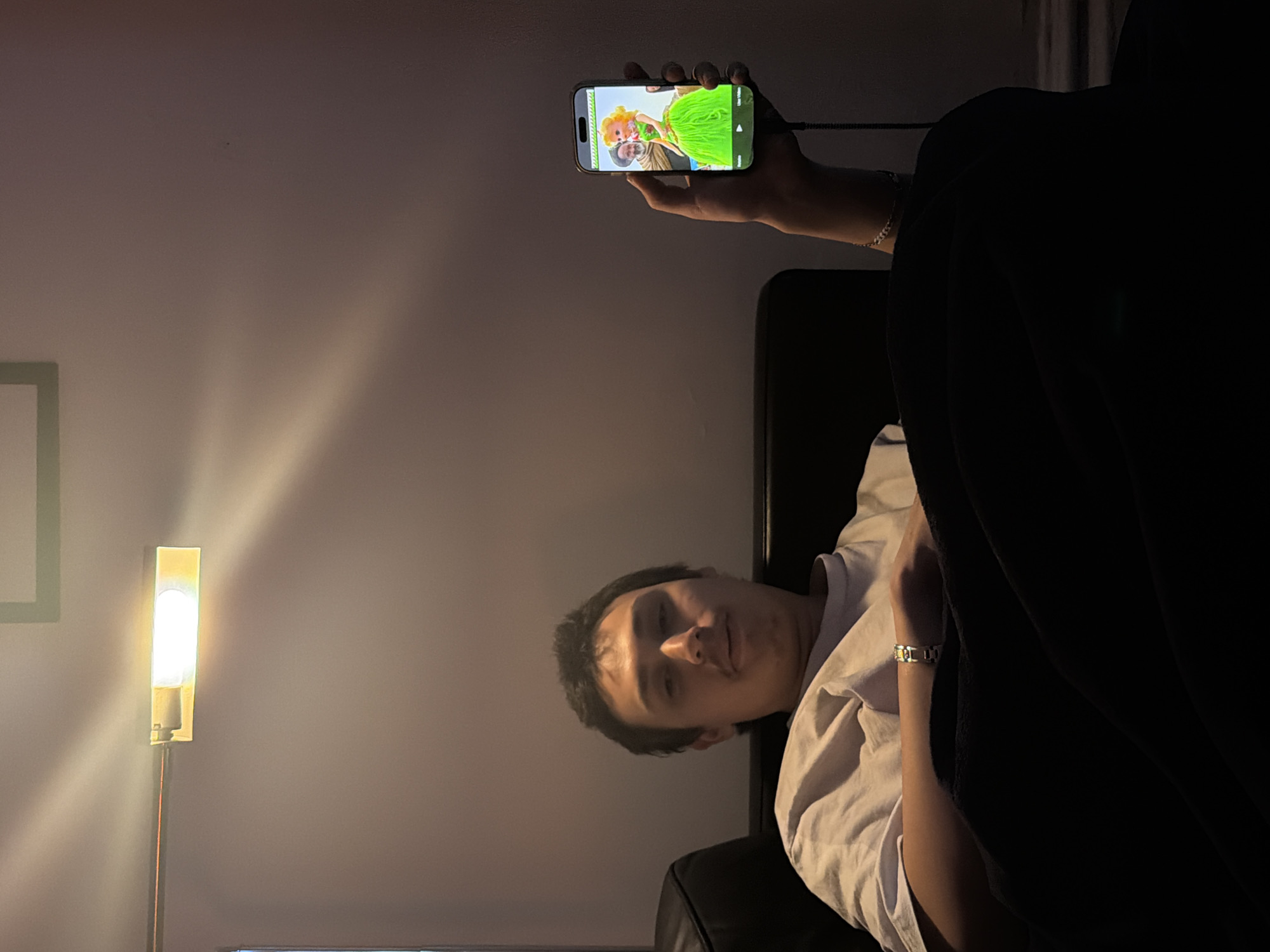 A person lies on a black leather sofa, illuminated by a floor lamp. They are holding a smartphone, displaying a video. The background is a plain wall, with an outline of a frame. The location is indeterminate, and there is no evidence to suggest it is in New York City.