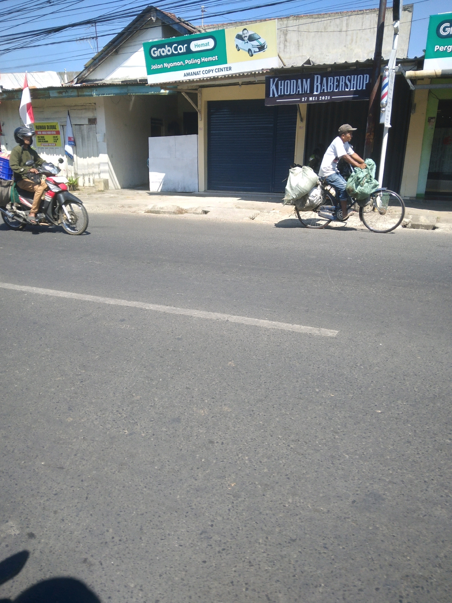 In Bancarkembar, Indonesia, a man is riding a bicycle laden with two large, green, cloth-covered bundles on a sunny day. The man wears a dark baseball cap, a white t-shirt, and blue jeans. The bicycle has a wire basket on the front and is equipped with fenders. Behind him, another person rides a motorcycle, wearing a dark helmet and a tan outfit. The motorcycle is red and black, with a blue crate attached to the rear. Both individuals are moving along a paved road. In the background, a strip of buildings lines the street, featuring various signs, including one that reads "GrabCar Hemat" and another that says "KHODAM BABERSHOP". The sky is clear and blue, and the sun casts shadows on the road.