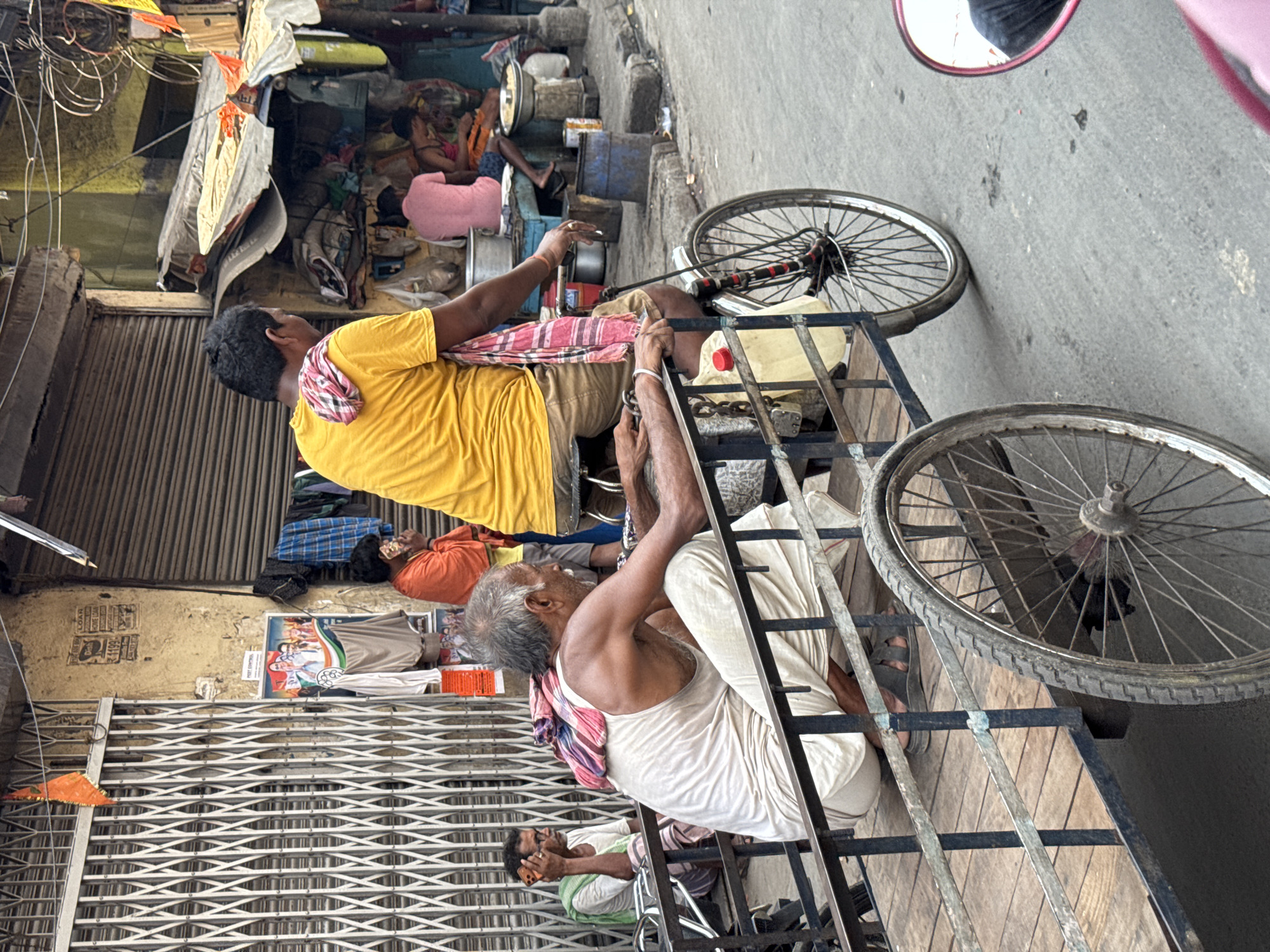 The image captures a street scene in Kolkata, India. Two men are on a rickshaw. One man, wearing a yellow shirt, is on the rickshaw, while the other, wearing a white outfit, is pulling it. The road is asphalt. Buildings line the street, one with a closed metal shutter. People are resting and sleeping near the building. Wires are above.