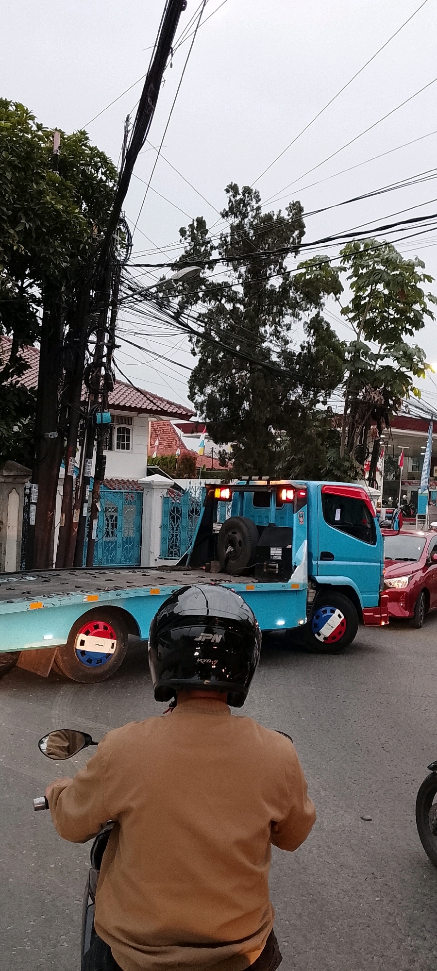 In Depok, Indonesia, a light blue tow truck is positioned on an asphalt road, its rear platform lowered, carrying a spare tire. A red sedan is stopped behind the tow truck. In the foreground, a person wearing a brown jacket and a black helmet is seated on a motorcycle, facing away from the camera. Overhead, a dense network of power lines crisscrosses the cloudy sky. In the background, a white house with a red tiled roof is visible, partially obscured by trees and foliage. The scene captures a street view with vehicles, infrastructure, and residential elements under an overcast sky.