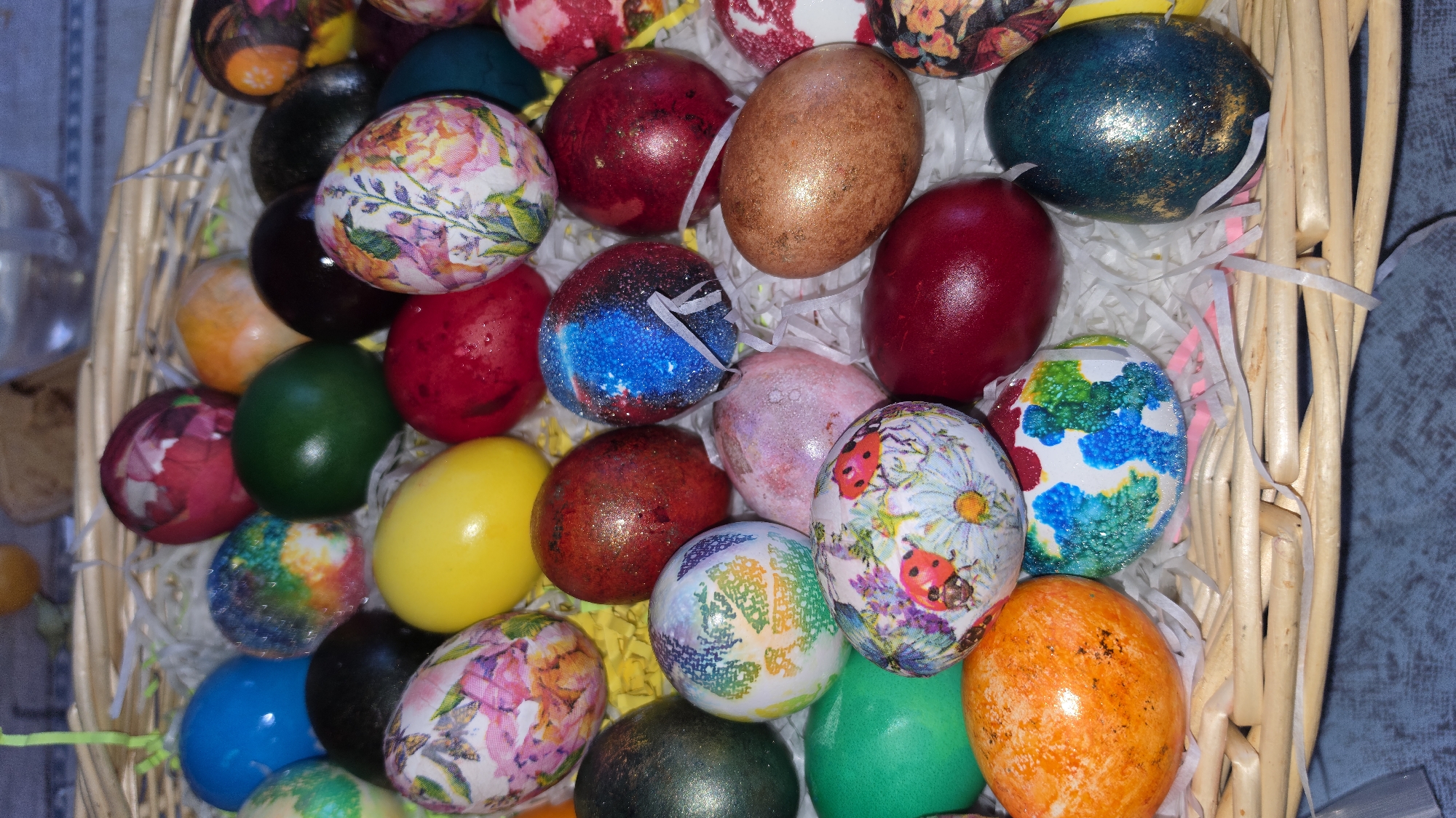 A close-up, eye-level shot depicts a woven basket filled with a variety of decorated Easter eggs nestled in white shredded paper. The eggs are multi-colored and feature diverse patterns and designs, including solid colors, floral motifs, and textured surfaces. Some eggs appear to have glitter or other decorative elements applied. The background consists of a neutral-toned fabric or surface. There are no identifiable people present in the frame. The image does not contain information related to the city of London, United Kingdom.