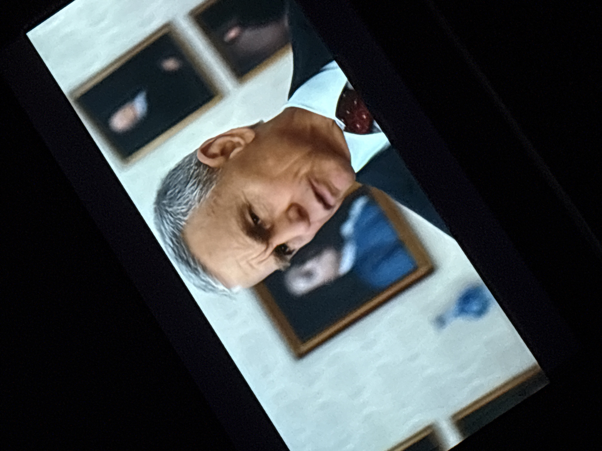 A man with gray hair and a suit is looking to the right. He is in a room with paintings on the wall. One painting shows a person in a blue shirt. There is also a blue vase on the wall. The image appears to be a screenshot from a movie or television show, and the location is not identifiable.