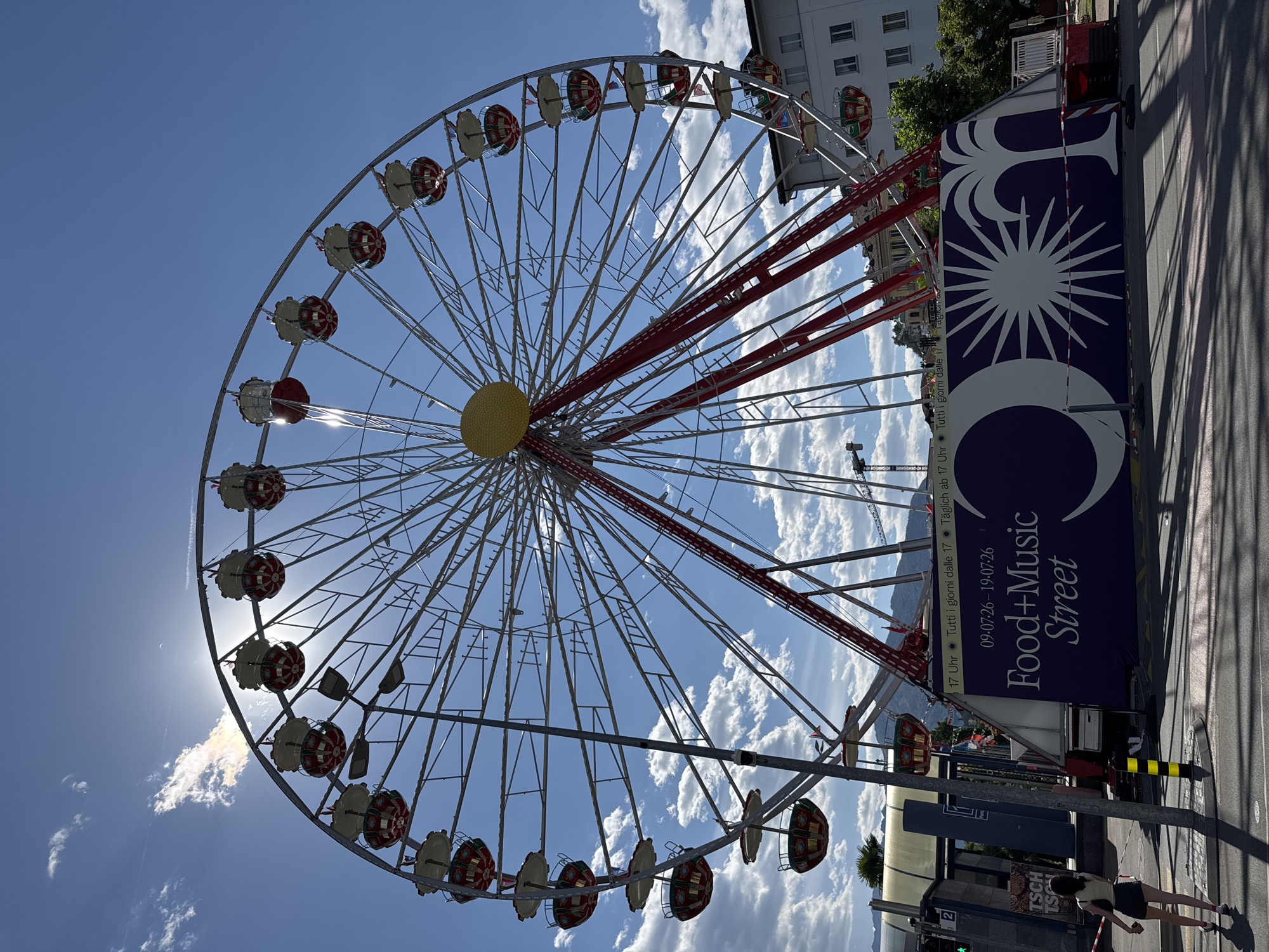 This is a media file depicting a Ferris wheel at an outdoor event. The Ferris wheel is large and appears to be white with red accents on its spokes and gondolas, which are circular and red and white. The wheel is seen from a low angle, looking up towards the blue sky with scattered white clouds. The sun is bright and is partially obscured by the wheel, creating a lens flare.

On the right side of the image, there is a large, dark blue banner with white text and graphics. The text reads "Food+Music Street" and provides dates and times for the event: "09.07.26-19.07.26" and "17 Uhr Tutti i giorni dalle 17 Täglich ab 17 Uhr Tutti i giorni dalle Tagicia". The graphics on the banner include a stylized sun and a crescent moon.

In the foreground, on the right, a street is visible with a yellow and black striped bollard and some shadows cast by the Ferris wheel and other structures. Two people are partially visible on the sidewalk, with one woman in a white top and dark skirt walking away from the camera. The presence of the banner advertising a "Food+Music Street" event and the Ferris wheel suggests a fair, festival, or amusement park setting. The bright sunlight and blue sky indicate it is daytime, likely a sunny day. The location context provided, Locarno, Switzerland, adds to the understanding of the setting.