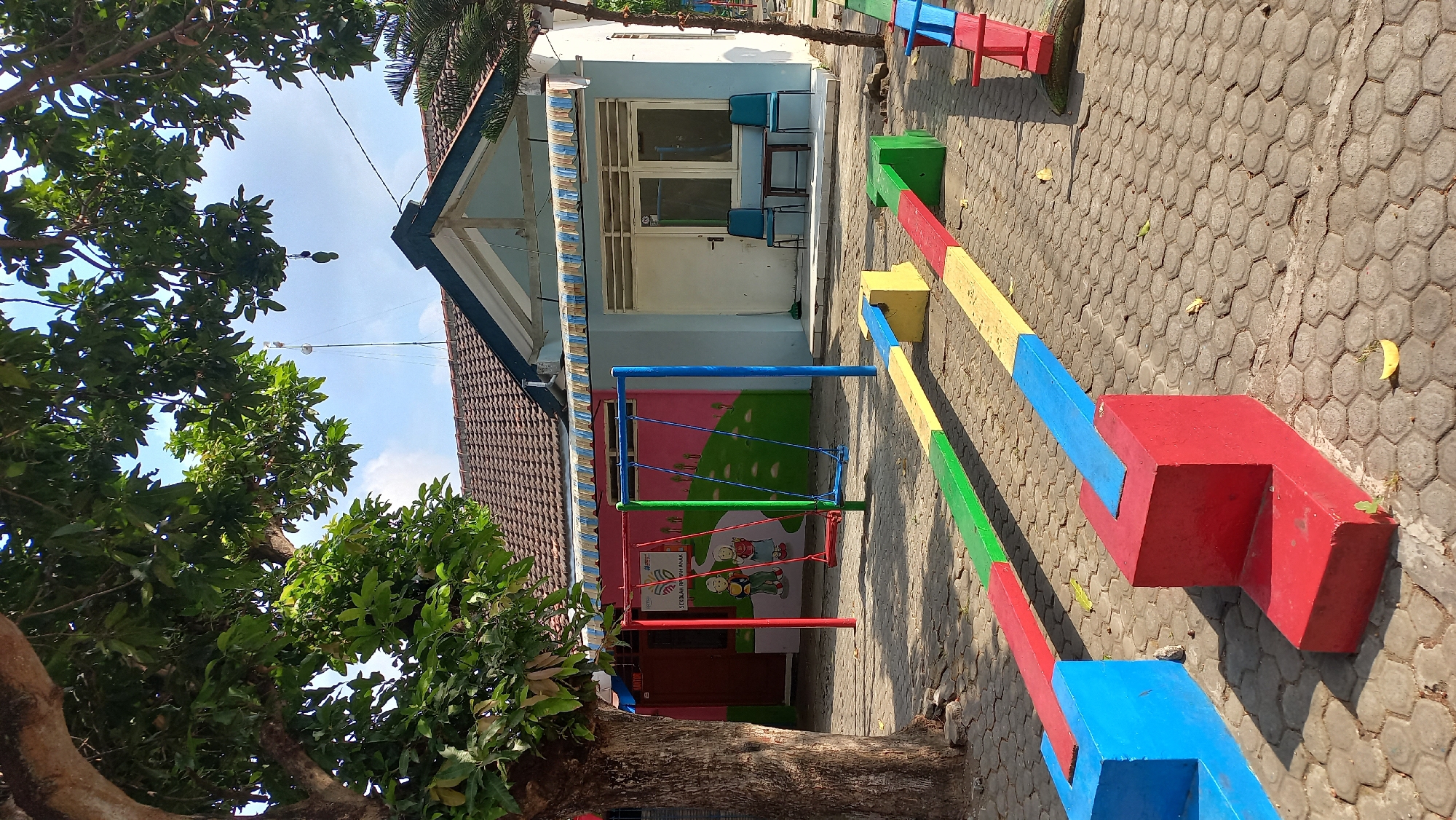 In the city of Kenayan, Indonesia, a sunlit outdoor scene depicts a children's play area in front of a light blue building with a brown tiled roof. A large tree with abundant green leaves shades the left side of the frame. In the foreground, a colorful seesaw-like play structure made of red, yellow, and green blocks is laid out on a paved hexagonal stone surface. To the left, a bright pink wall features a mural of two children holding flowers, accompanied by text indicating it is a "Sekolah PAUD Anak" (Children's Early Childhood School). A swing set with red, green, and blue poles and swings stands near this wall. The building behind the play area has a light blue awning over a section with two windows. Two blue chairs and a small dark table are placed on a small concrete porch in front of the building's entrance, which features a white door. The overall environment appears calm and suitable for children's activities.