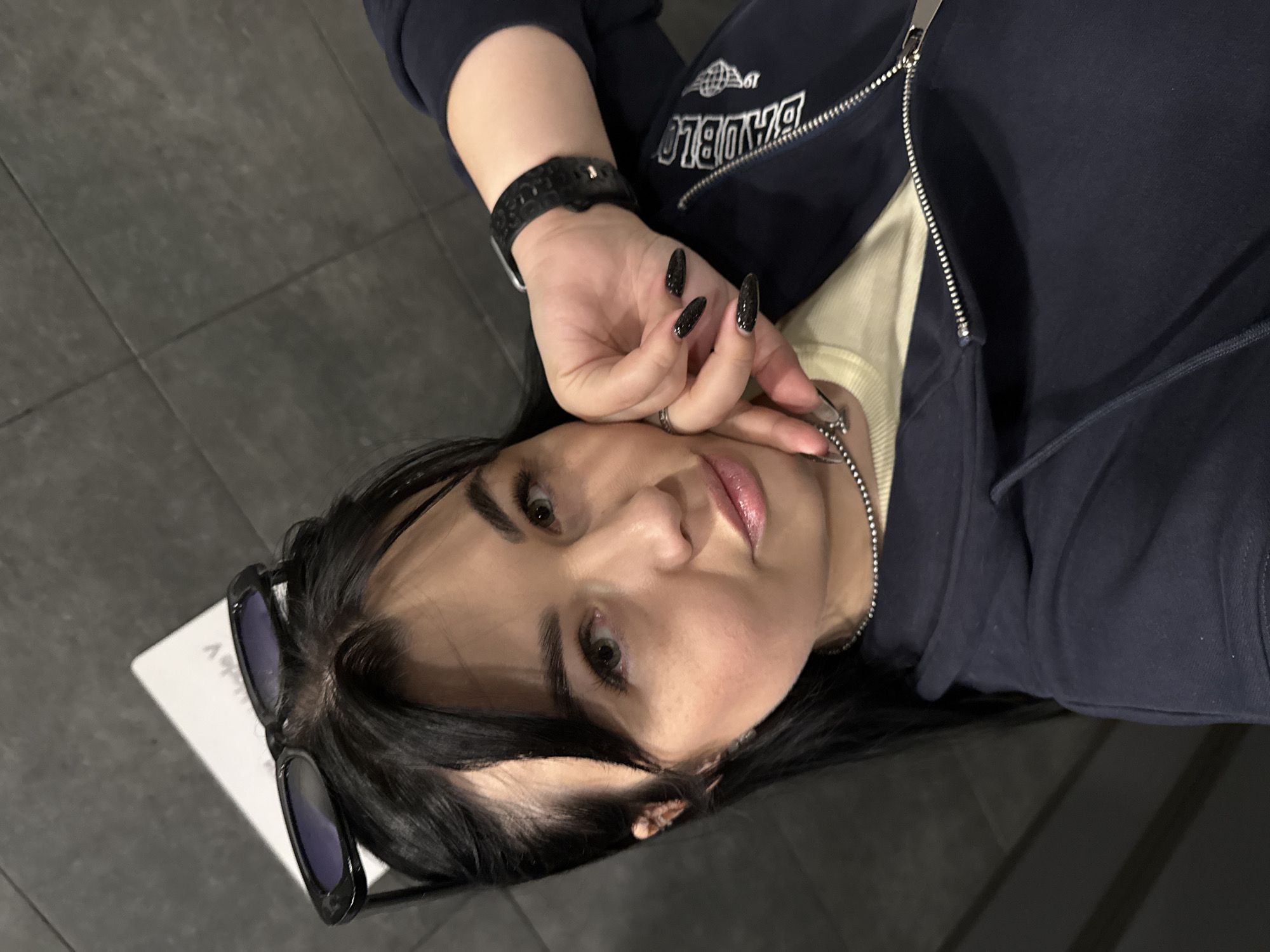 A medium close-up, rotated photograph shows a young woman lying on a dark tiled surface, looking directly at the camera. Her expression is calm with slightly parted lips. She has dark hair, and black sunglasses are perched on top of her head.
She is dressed in a dark blue zip-up hoodie, worn over a light yellow or cream-colored ribbed top. A silver chain necklace is visible around her neck, with her right hand gently touching it. This hand displays several rings and long, pointed nails painted with dark, glittery polish. A black smartwatch is worn on her left wrist. On the sleeve of her hoodie, a stylized "UMBRO" logo is visible, along with text that appears to be "BANDBL0" or a similar mirrored/stylized brand name.
The setting appears to be indoors, indicated by the dark tiled floor and uniform lighting. A small portion of a white object, possibly a book or tablet, is visible behind her head. The activity is a posed portrait or selfie. The location is Perm, Russia.