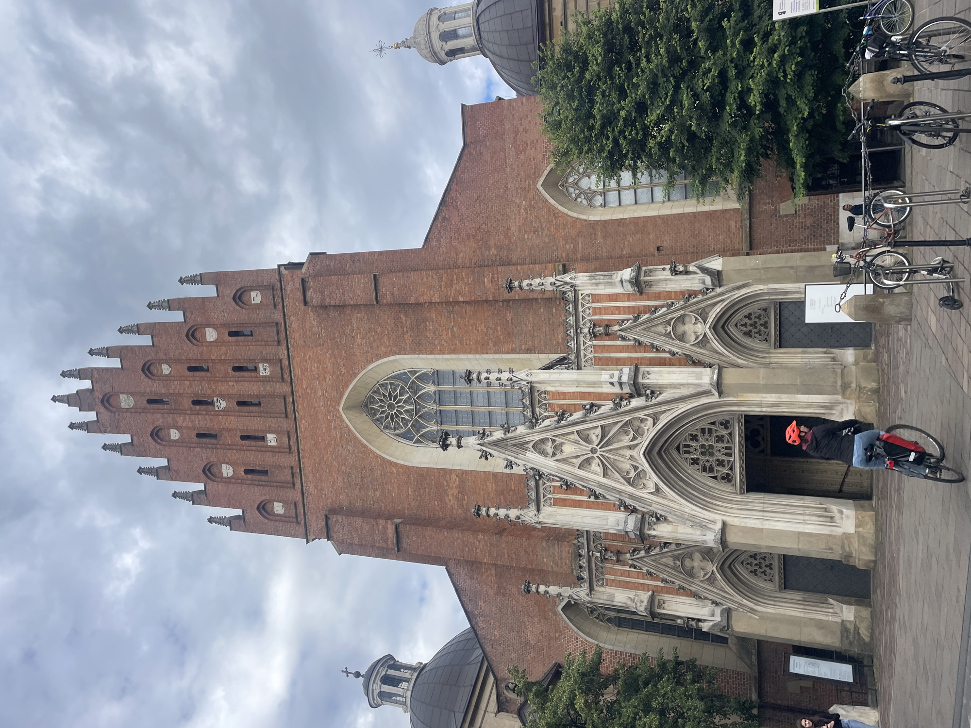 The image shows a wide-angle, eye-level view of the facade of St. Mary's Basilica in Krakow, Poland, on a cloudy day. The grand Gothic brick structure dominates the frame, with its intricate spires and arched windows. In the foreground, a person wearing a red helmet is riding a bicycle away from the camera. To the left of the entrance, a woman stands near a sign. Bicycles are parked on the right side of the plaza. Lush green trees flank the sides of the basilica, and a large, dark dome with a smaller, lighter dome on top is visible on the left side. The cloudy sky provides diffused lighting for the scene.