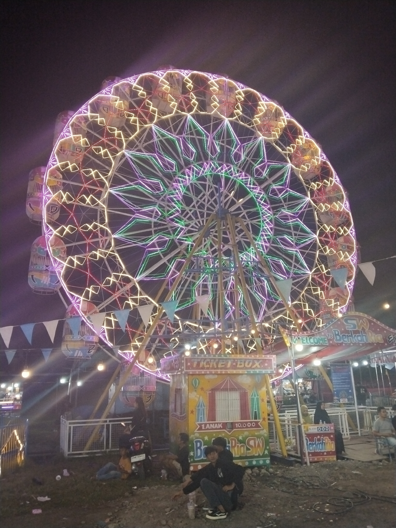 The image depicts a night scene at a fairground in Sragen, Indonesia. The focal point is a large Ferris wheel illuminated with multicolored lights, primarily pink, green, yellow, and white. Below the Ferris wheel, a ticket box is visible, painted with bright colors and the words "TICKET BOX" and "Berkah Ria SW." Several people are present in the lower portion of the image, some sitting on the ground near the ticket box, and others standing or sitting nearby. A parked motorcycle is also visible near the ticket box. String lights and pennant banners are strung across the fairground, adding to the festive atmosphere. The ground appears to be a mix of grass and dirt. In the background, additional fairground structures and people can be seen, though less clearly.