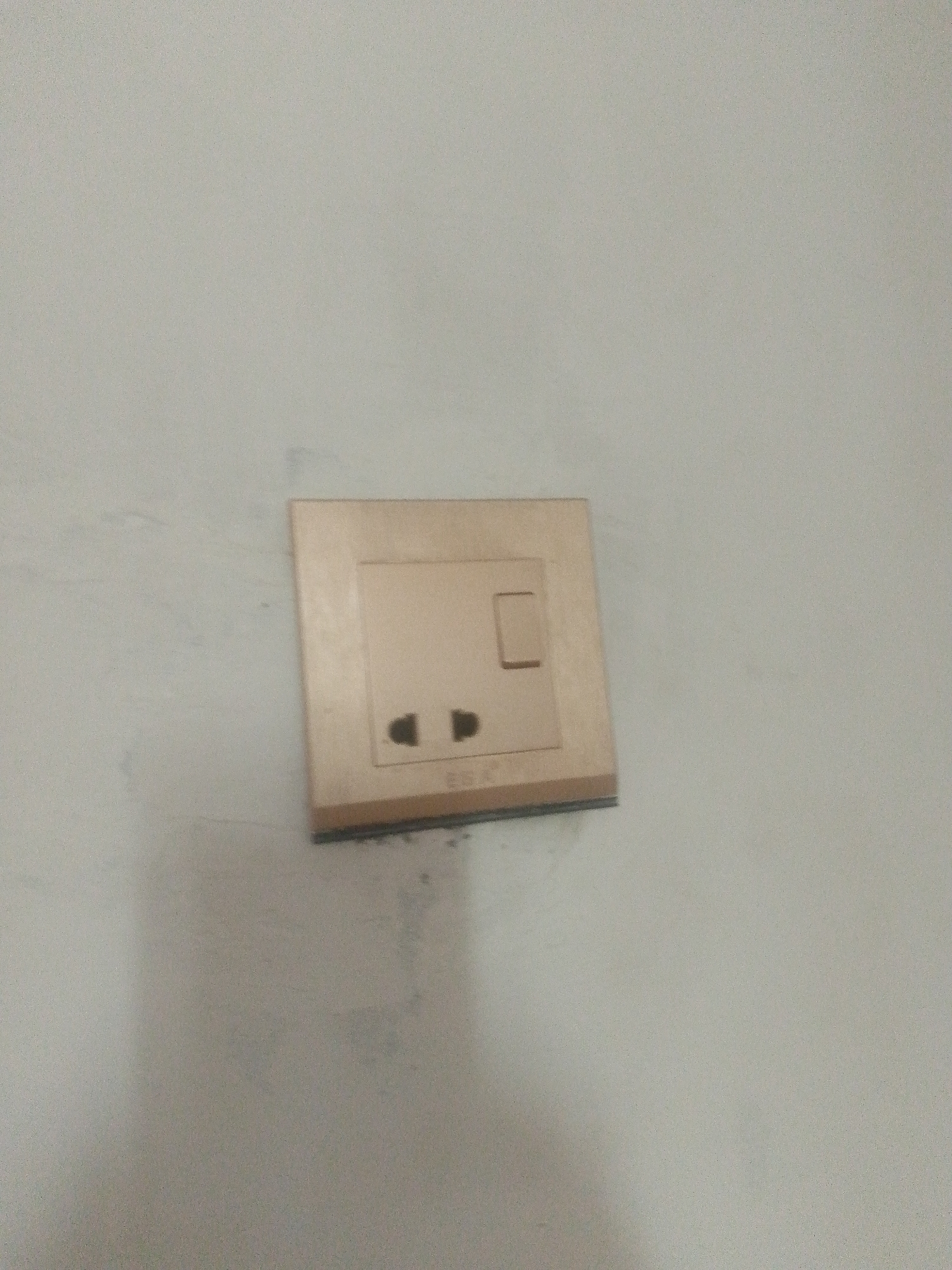 The image shows a close-up of a beige electrical outlet with a switch, mounted on a light-colored, textured wall. The outlet has two circular openings and a rectangular switch located above them. The overall lighting is dim, creating shadows and a slightly grainy texture. The scene appears to be indoors, and without further information or context, it is impossible to determine its specific location within the city of Surahani, Azerbaijan. The image is static and depicts a common household fixture.