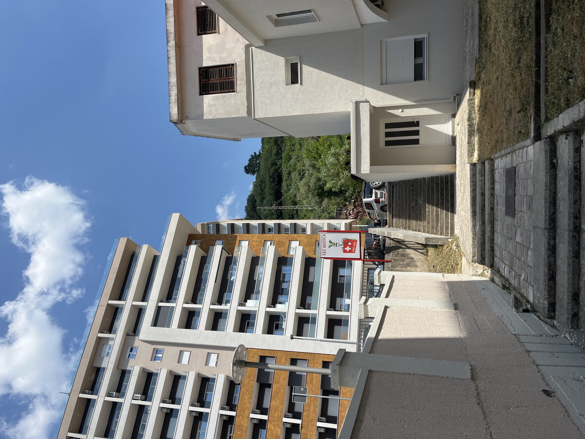 In Budva, Montenegro, a modern apartment building with a beige and brown facade stands tall under a bright blue sky with scattered clouds. To its right, a smaller, older white building features dark-shuttered windows. A red sign with a white cross and the words "VET MEDICA FIRST AID" is prominently displayed between the two buildings. A paved pathway leads up a set of stone stairs towards the white building, with a silver SUV parked at the top. The surrounding area is characterized by lush green hillsides and a dry, grassy patch of ground.