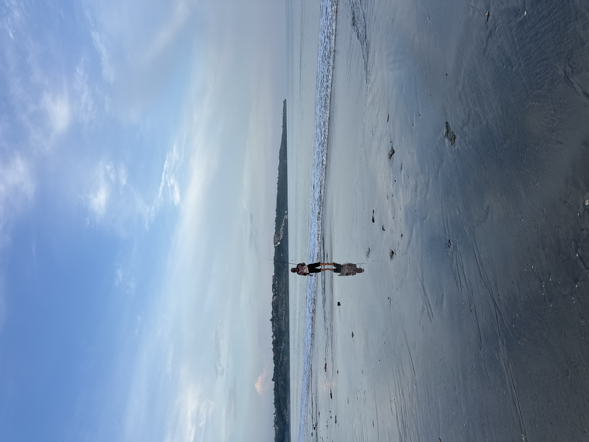 The image shows a beach scene in Jimbaran, Indonesia. A man stands on the wet sand near the shoreline, holding a fishing rod. The tide is coming in, with small waves visible. A lush, forested shoreline is located behind the man. The sky is blue with scattered clouds.