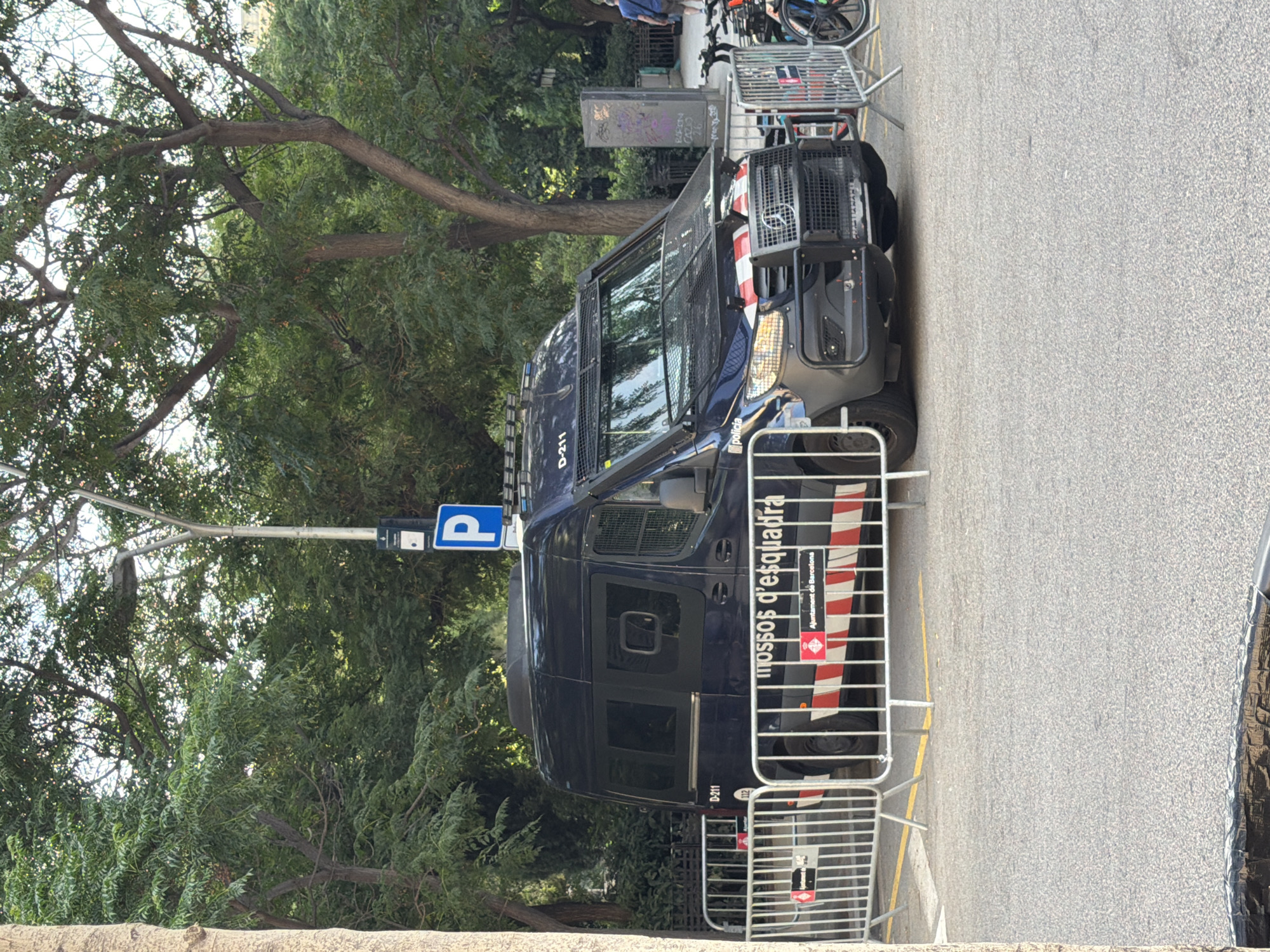 In Barcelona, Spain, a dark blue police van, identified by the markings "Mossos d'Esquadra" and "Ajuntament de Barcelona", is parked on an asphalt surface, cordoned off by temporary silver metal barriers. The van, equipped with a reinforced front grille and flashing lights, has the designation "D-211" visible on its upper front. A blue sign with a white "P" indicates a parking area. Trees with dense green foliage form a backdrop to the scene, with a street light pole also present. In the background, a person in a blue shirt walks away from the camera, and bicycles are visible parked behind the barriers. The ground is marked with yellow lines.