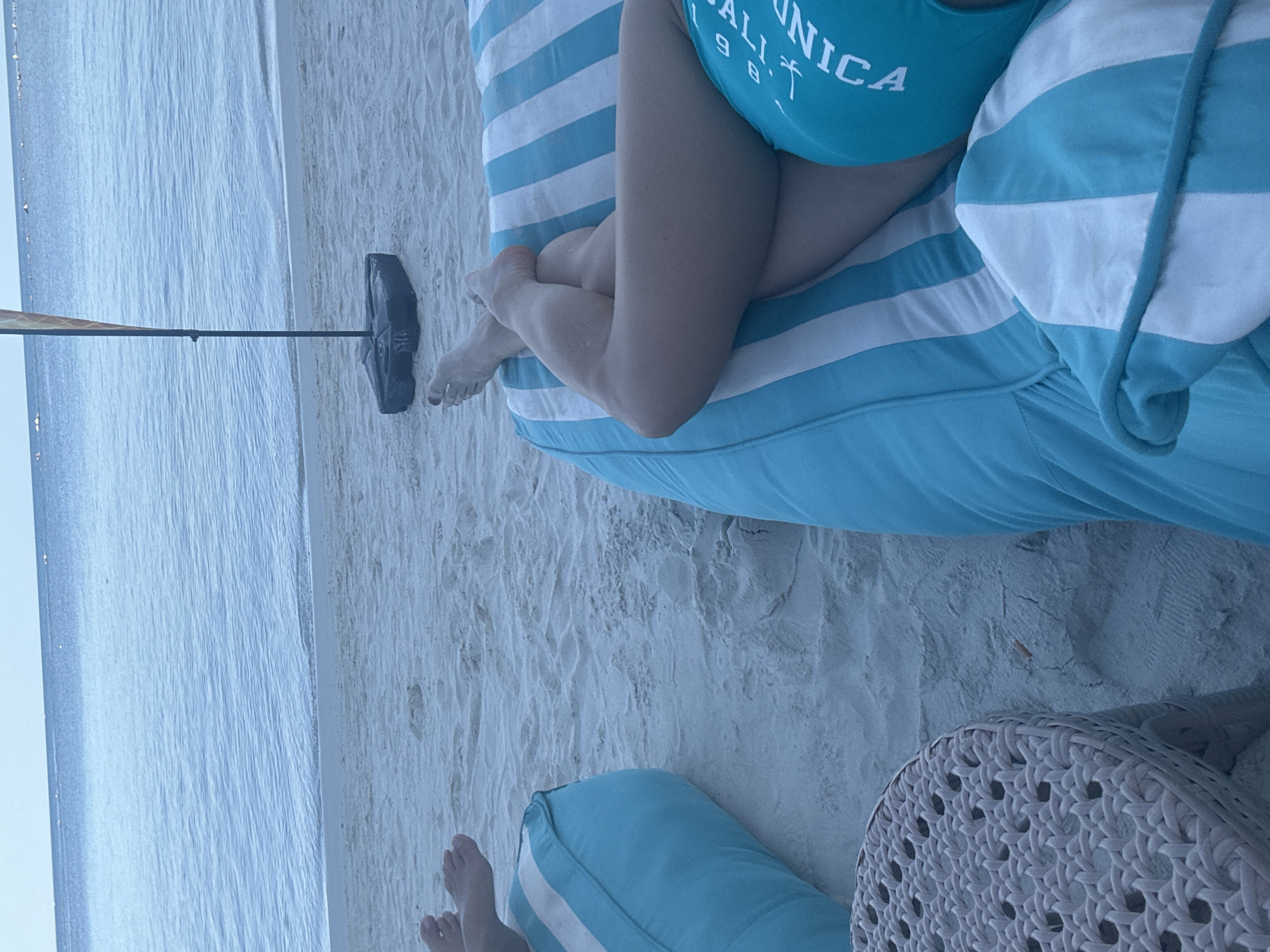 This is a medium shot, eye-level, outdoor, daylight photograph taken on a sandy beach in the Ko Samui District of Thailand. The foreground features a woven, light-gray side table to the left, partially obscuring a large, turquoise and white striped beanbag chair. A person's legs, clad in a turquoise swimsuit with white lettering that reads "CALIFORNIA," are crossed and resting on the beanbag. The person's feet, with red nail polish, are visible at the top of the beanbag. Further to the left, another turquoise and white striped beanbag is visible, with another person's feet resting on it.
In the midground, the sandy beach stretches towards the ocean. A black umbrella base with a partially unfurled, multi-colored umbrella stands in the sand between the beanbag chairs and the water. The ocean in the background is calm, with a gentle horizon line where it meets a pale blue sky. The water is a light blue hue, with subtle ripples on its surface. The overall impression is one of relaxation and a tropical vacation setting.