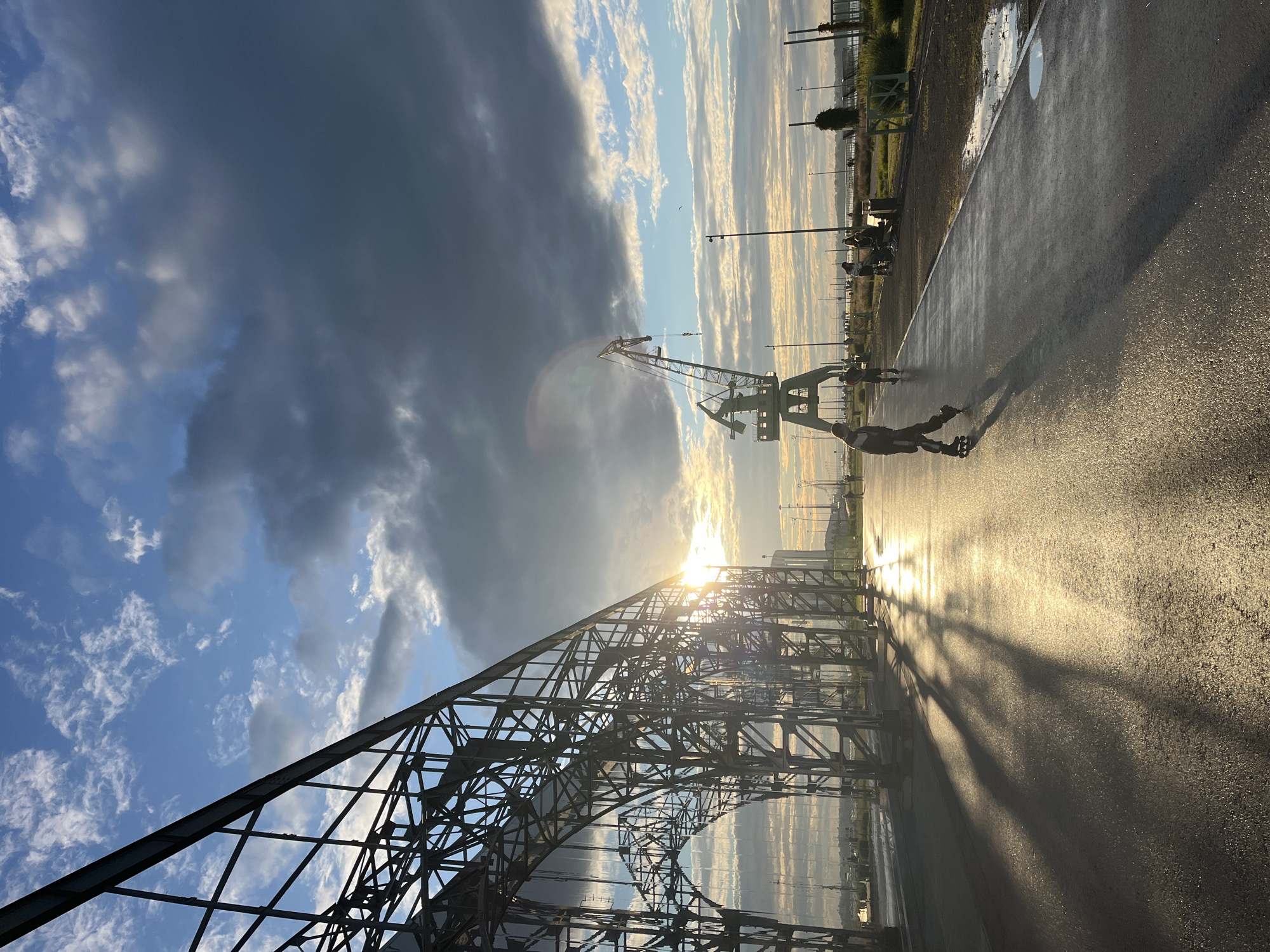 The image depicts an outdoor scene at sunset in Nizhny Novgorod, Russia. The foreground features a person rollerblading on a wet asphalt path, their silhouette cast in the golden light of the setting sun. Behind the rollerblader, a large, industrial crane stands tall against a dramatic sky filled with both dark, ominous clouds and bright, sunlit streaks. To the left, the skeletal structure of a large metal building or bridge stretches across the frame, its intricate latticework catching the light. In the distance, more industrial structures and vegetation are visible, along with a few other people scattered along the path, some sitting on a bench. The wet ground and puddles suggest recent rain, and the warm, low-angle sunlight indicates it is either sunrise or sunset, with the intensity and colors leaning towards sunset. The overall mood is one of urban industrial beauty blended with the dynamic atmosphere of a dramatic sky and the simple joy of outdoor recreation.